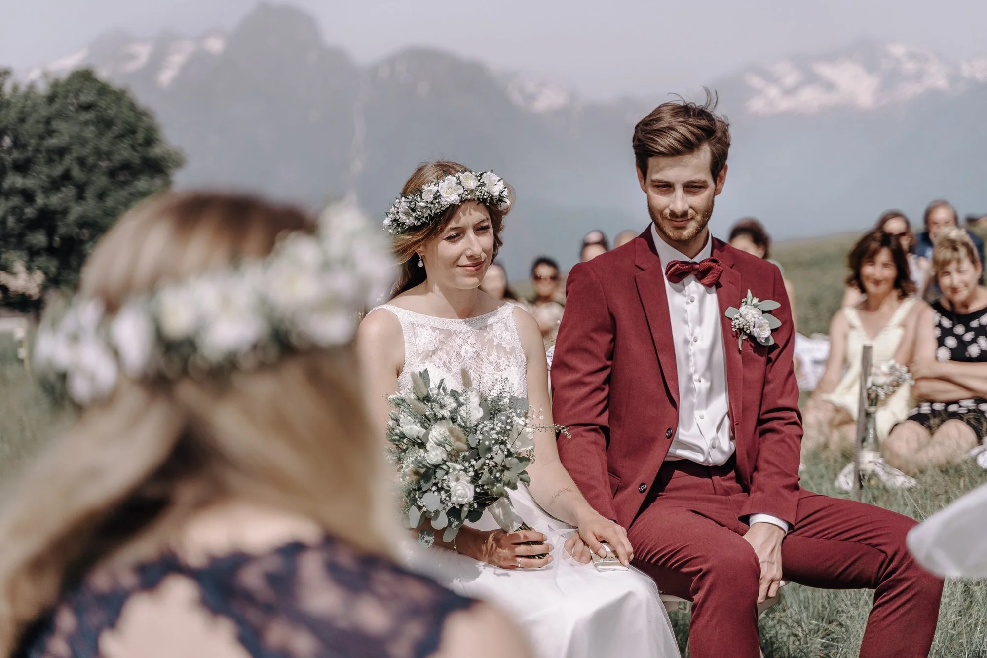 A young bride and groom sit side by side during their outdoor wedding ceremony, surrounded by guests in a scenic mountain setting. The bride wears a lace wedding dress and floral crown, while the groom wears a burgundy suit with a bow tie.