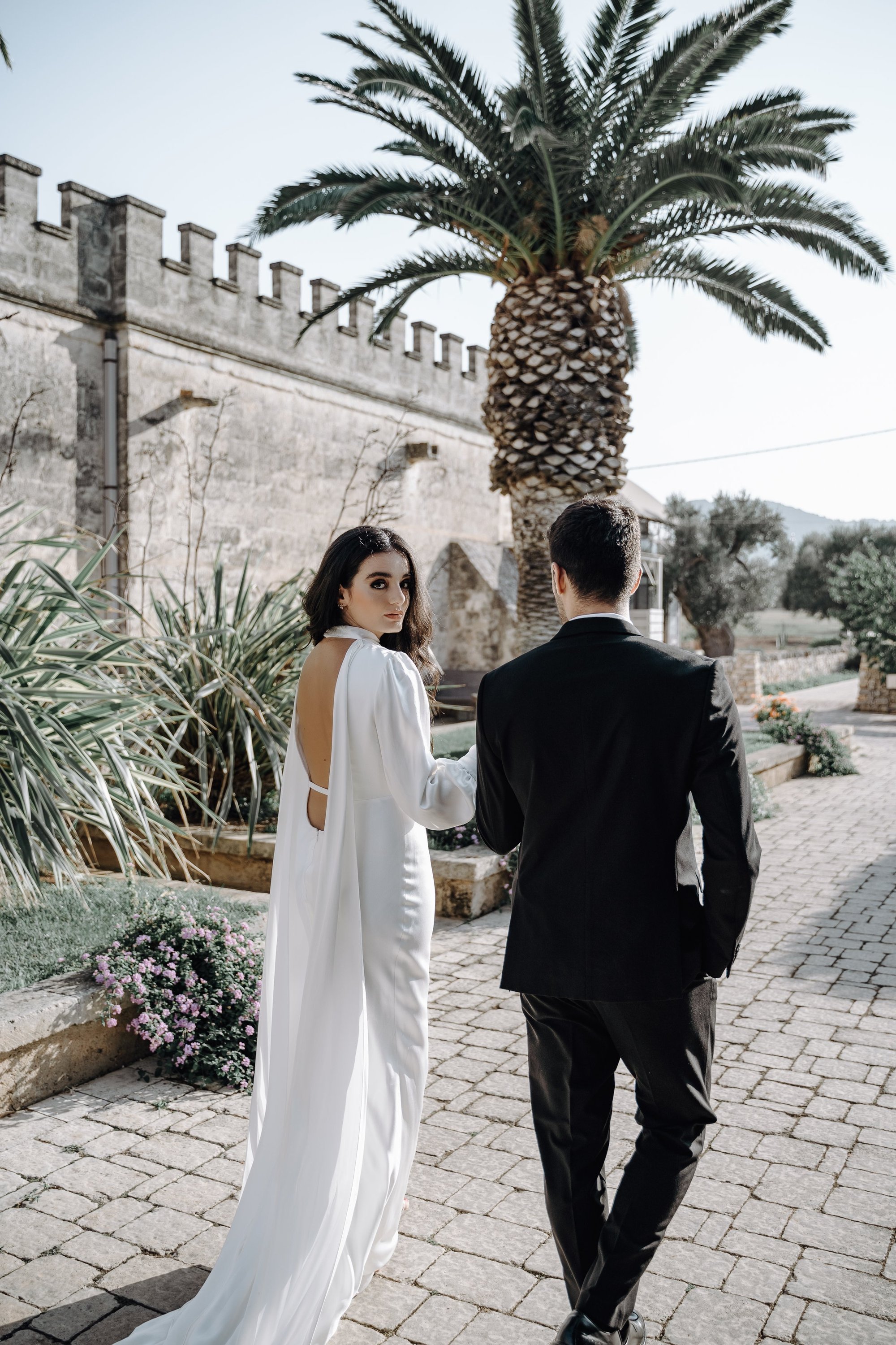 A woman in a white dress holding hands with a man in a black suit walking outdoors near a stone building with a large palm tree, flowers, and greenery.