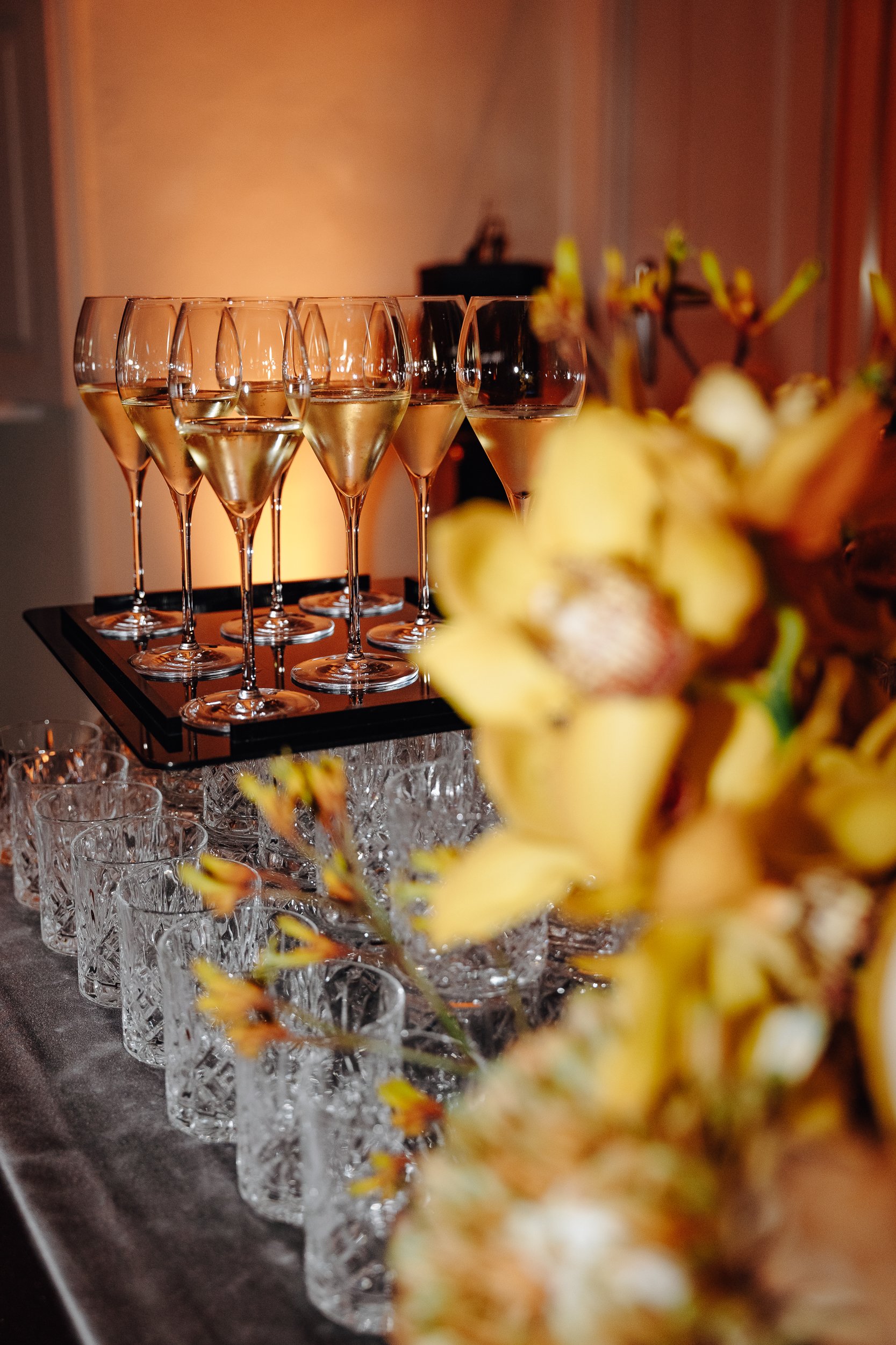 Several champagne glasses filled with sparkling wine arranged on a black tray at a celebration event with a blurred floral centerpiece in the foreground.