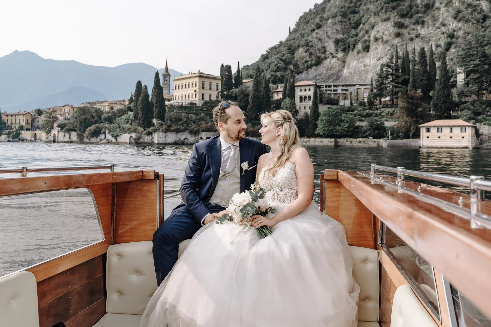 A newlywed couple sitting on a boat in a lake, with mountains and a lakeside town with tall cypress trees in the background.