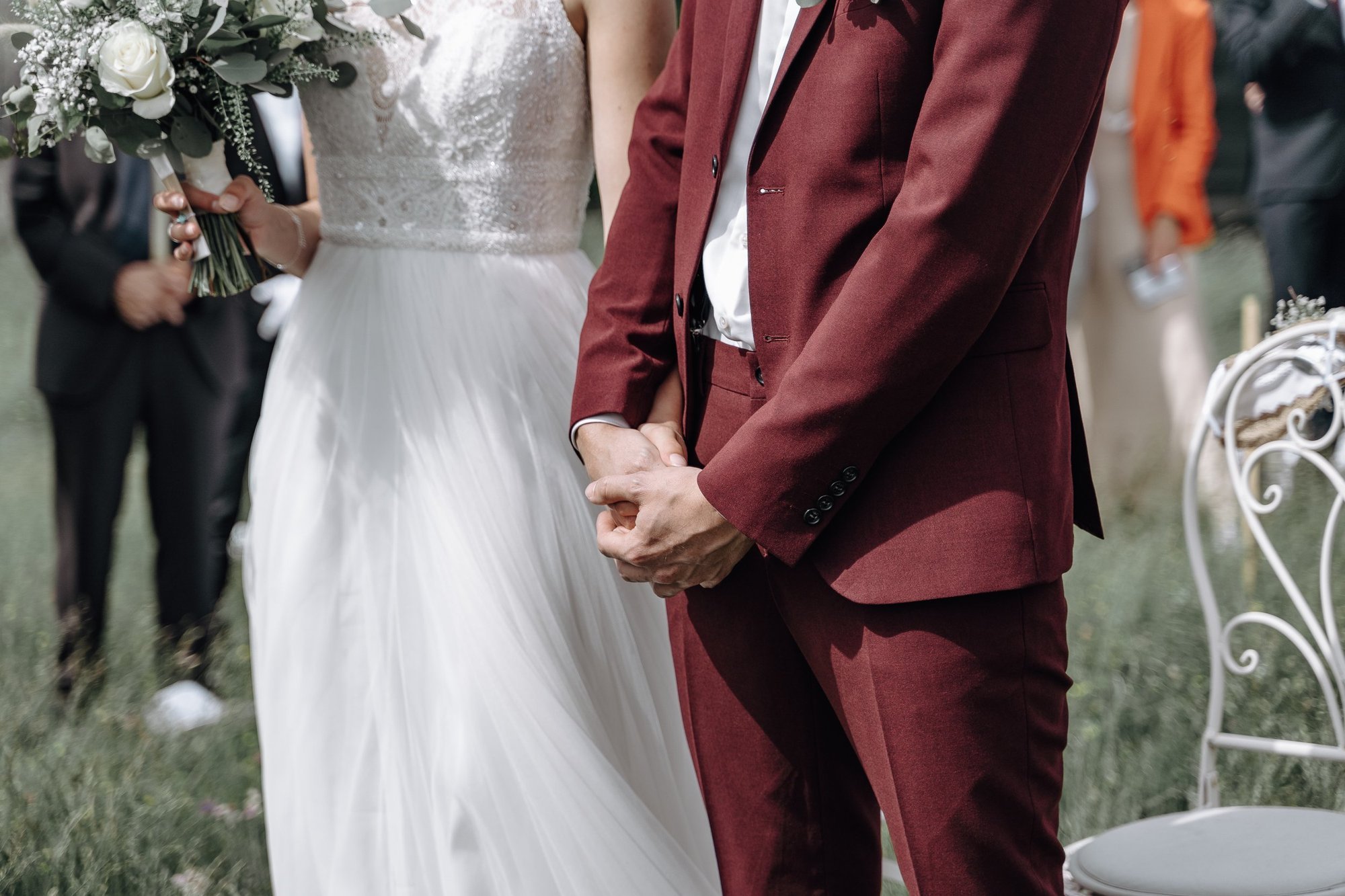 A bride and groom holding hands during a wedding ceremony outdoors. The bride is wearing a white lace dress and holding a bouquet, while the groom is dressed in a maroon suit.