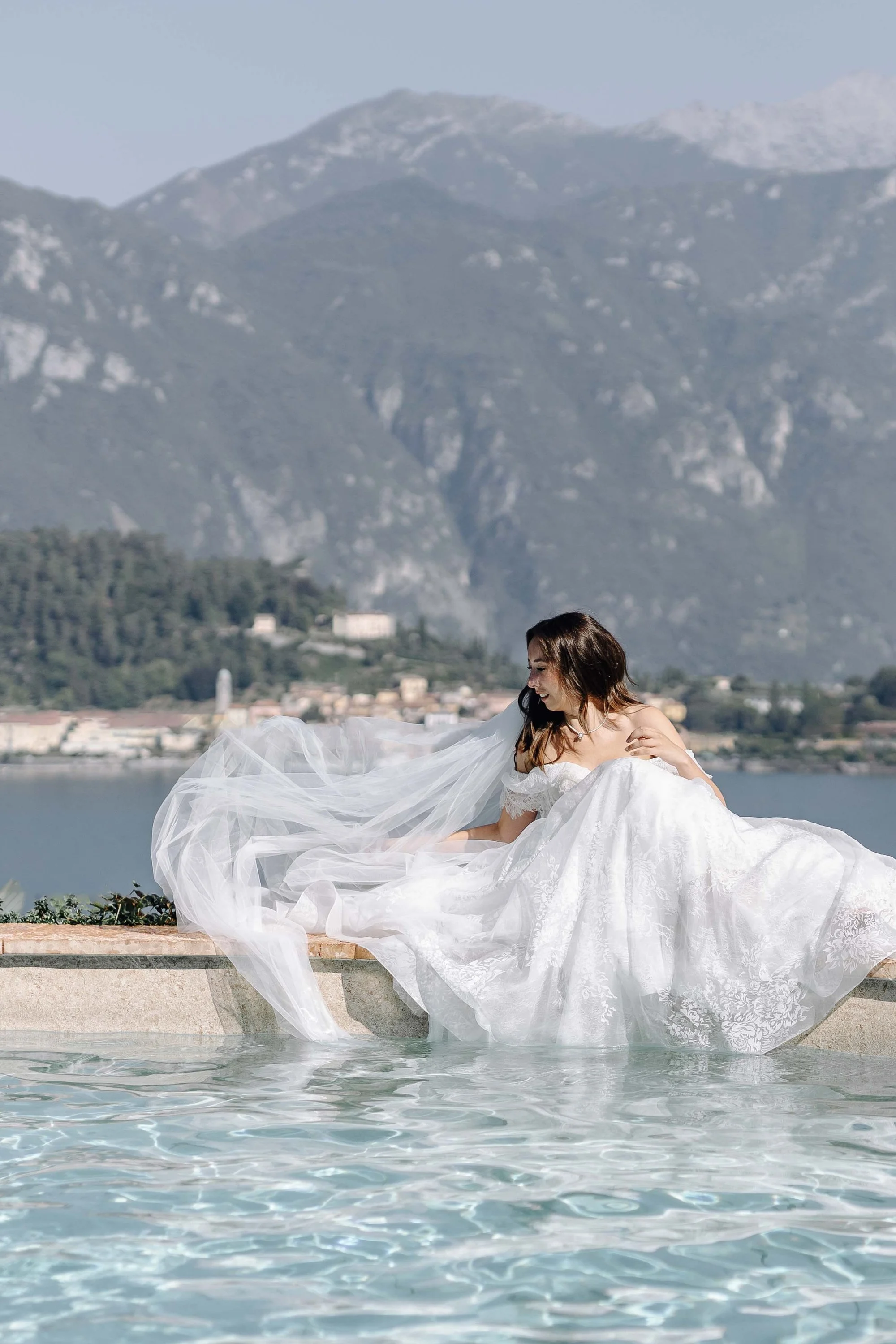 A woman in a white wedding dress sitting by a pool with a scenic mountain and town background.