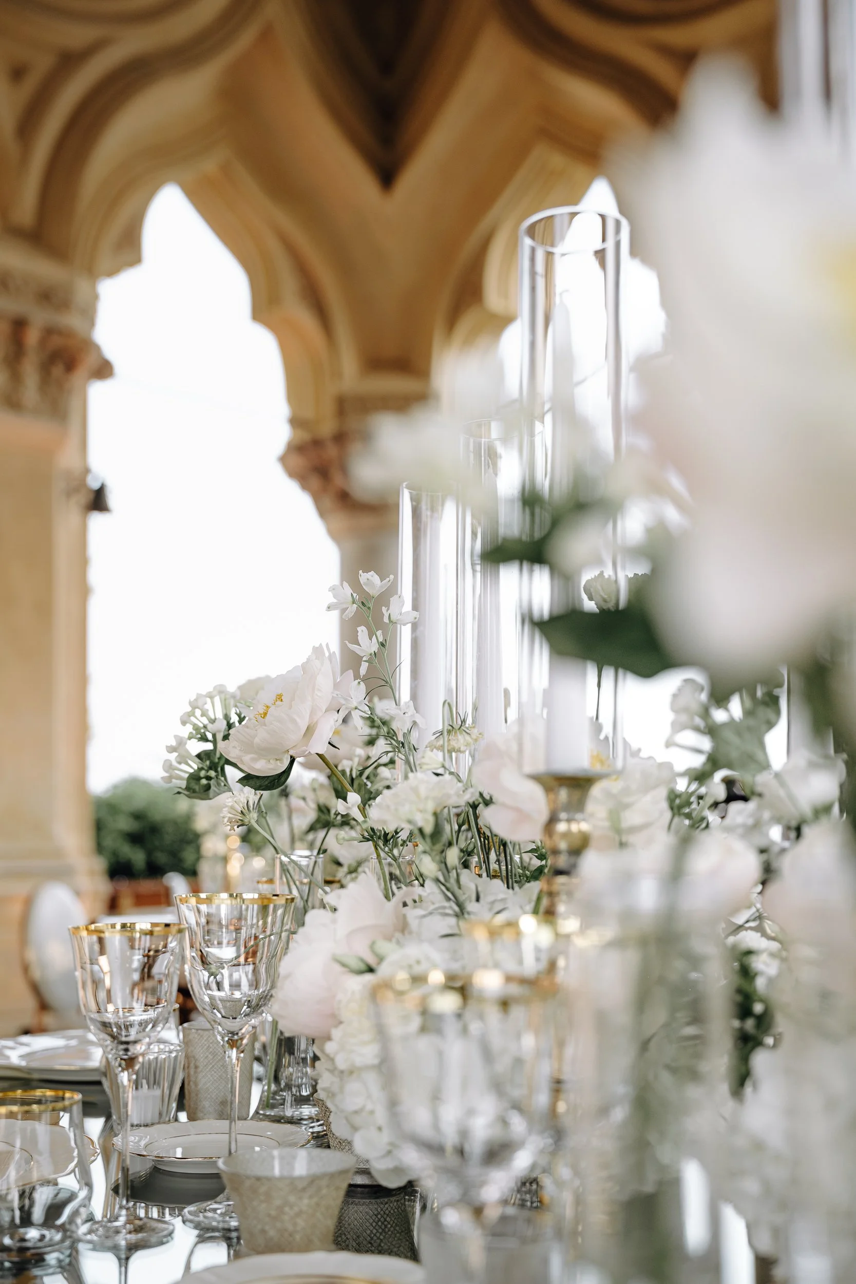 A decorated table with white flowers, glassware, and gold accents, set in an elegant indoor location with ornate architectural details on the ceiling.