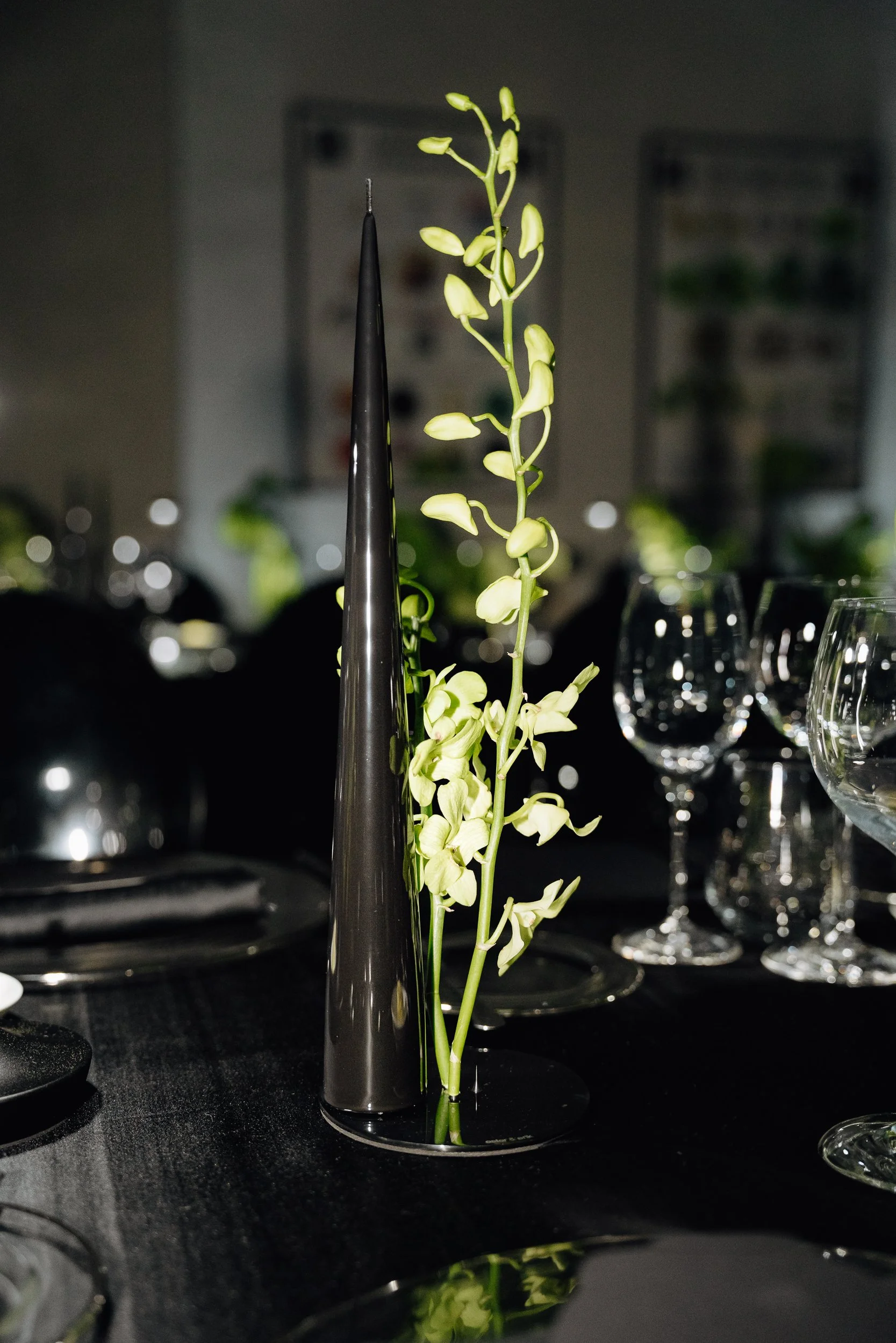 A tall, slender black vase with small yellow-green flowers inside. There are several wine glasses and dark plates on the table, with blurred background posters.
