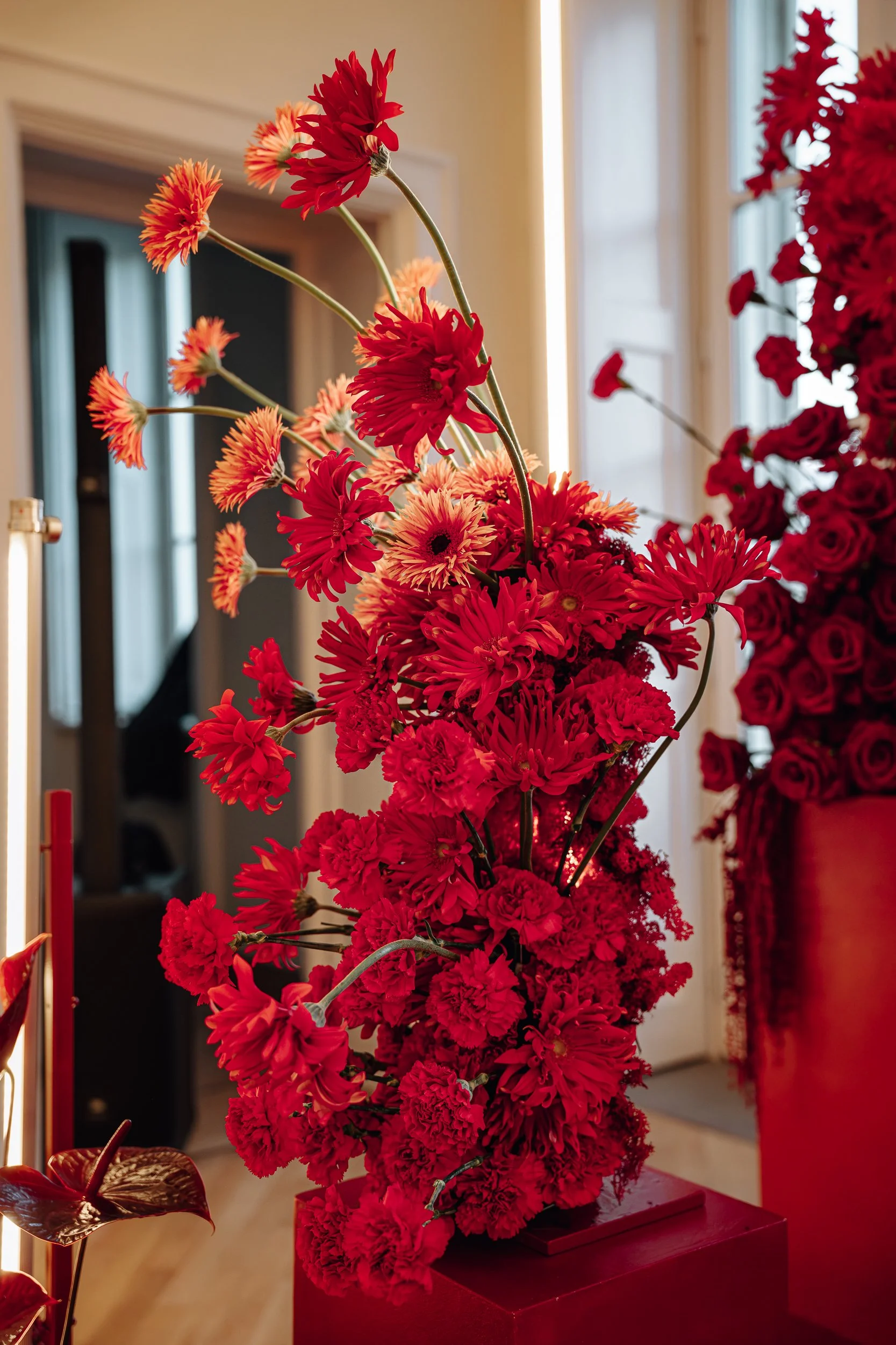 Close-up of a tall floral arrangement with red and orange flowers, displayed on a red pedestal in an indoor setting with beige walls and windows in the background.