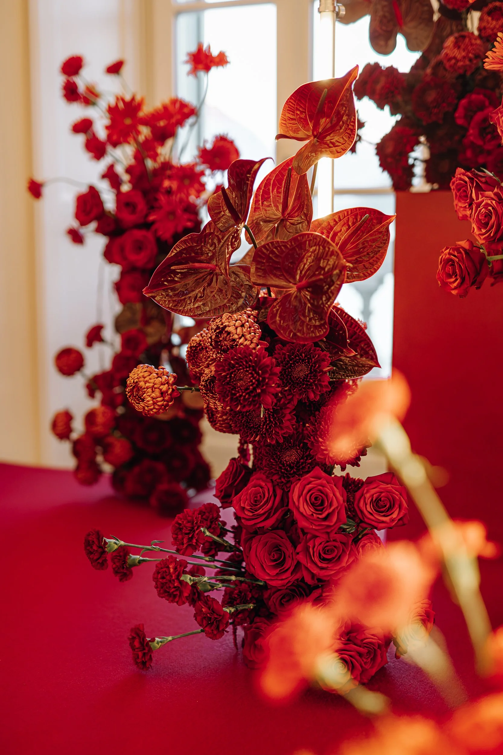 A floral arrangement featuring red roses, orange anthuriums, red carnations, and other red flowers, set on a red table with a blurred background.