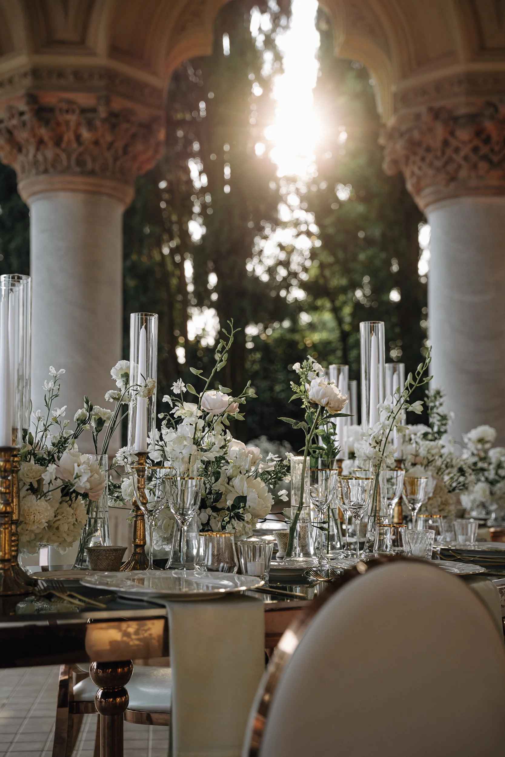 Elegant wedding or event table set with white flowers and tall glass candle holders, with sunlight streaming through architecture with columns and intricate carvings.