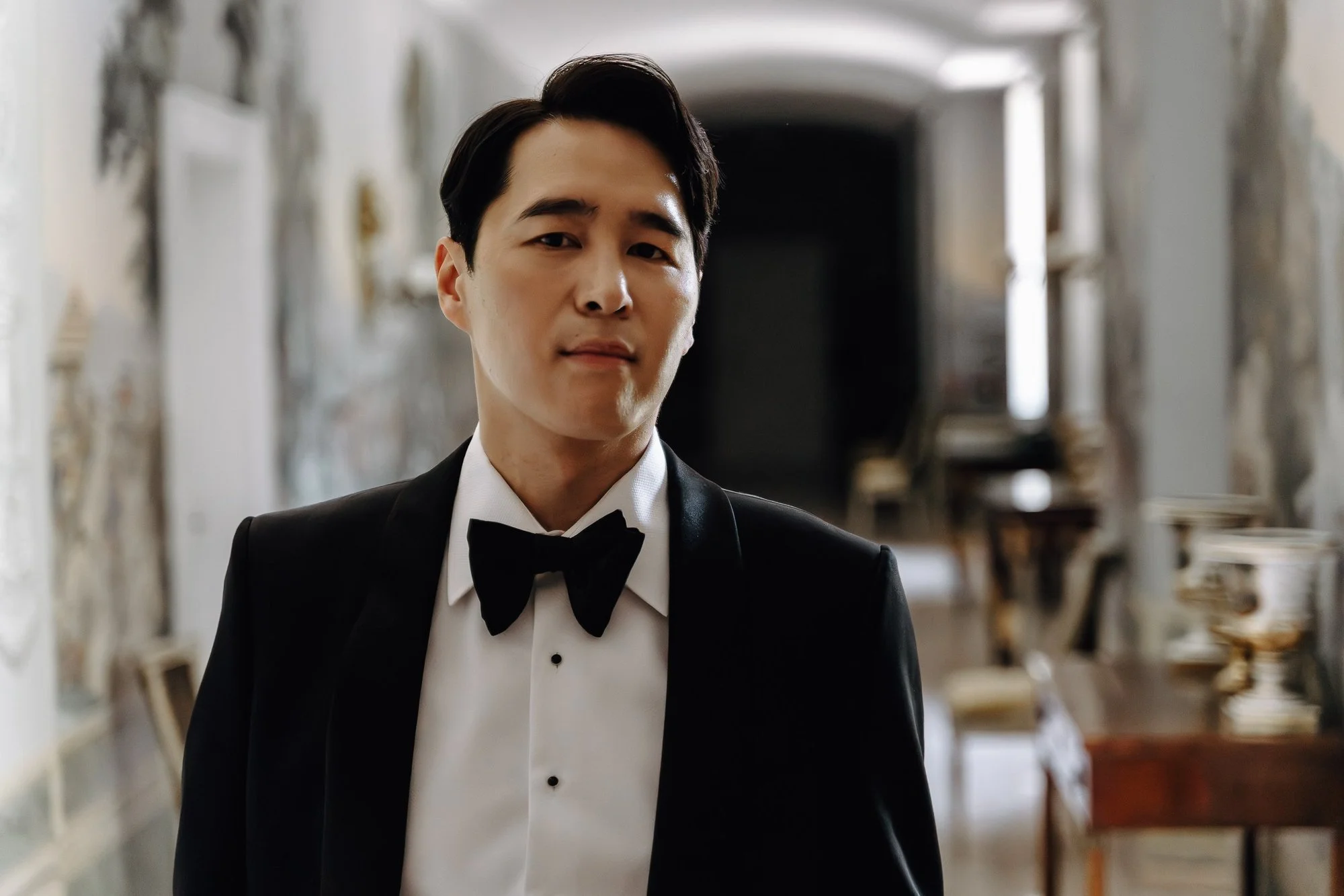A man wearing a black tuxedo and bow tie standing in a hallway with shelves and books in the background.