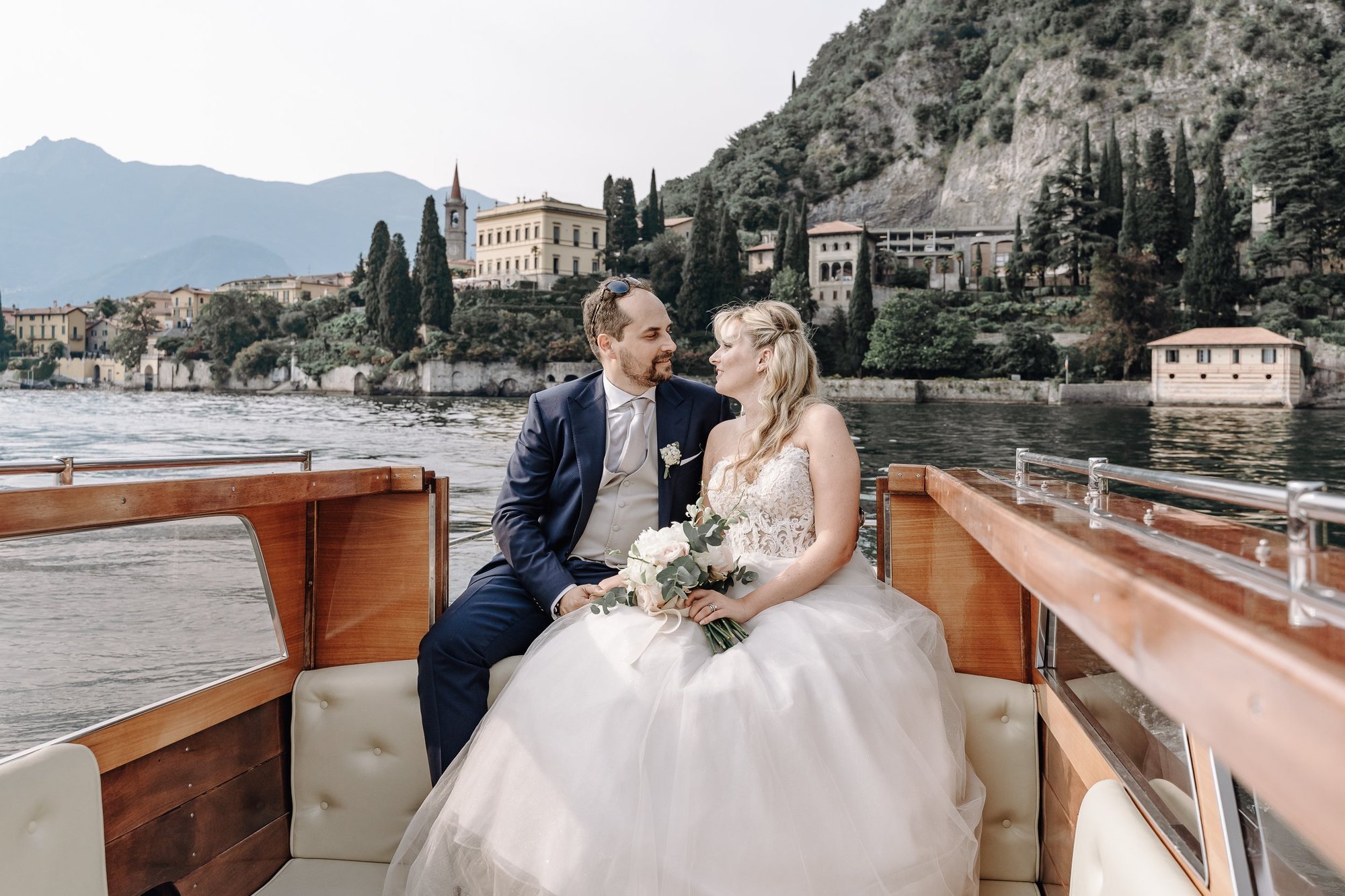 A bride and groom sitting closely together on a boat with mountains and a lakeside village in the background, during their wedding.