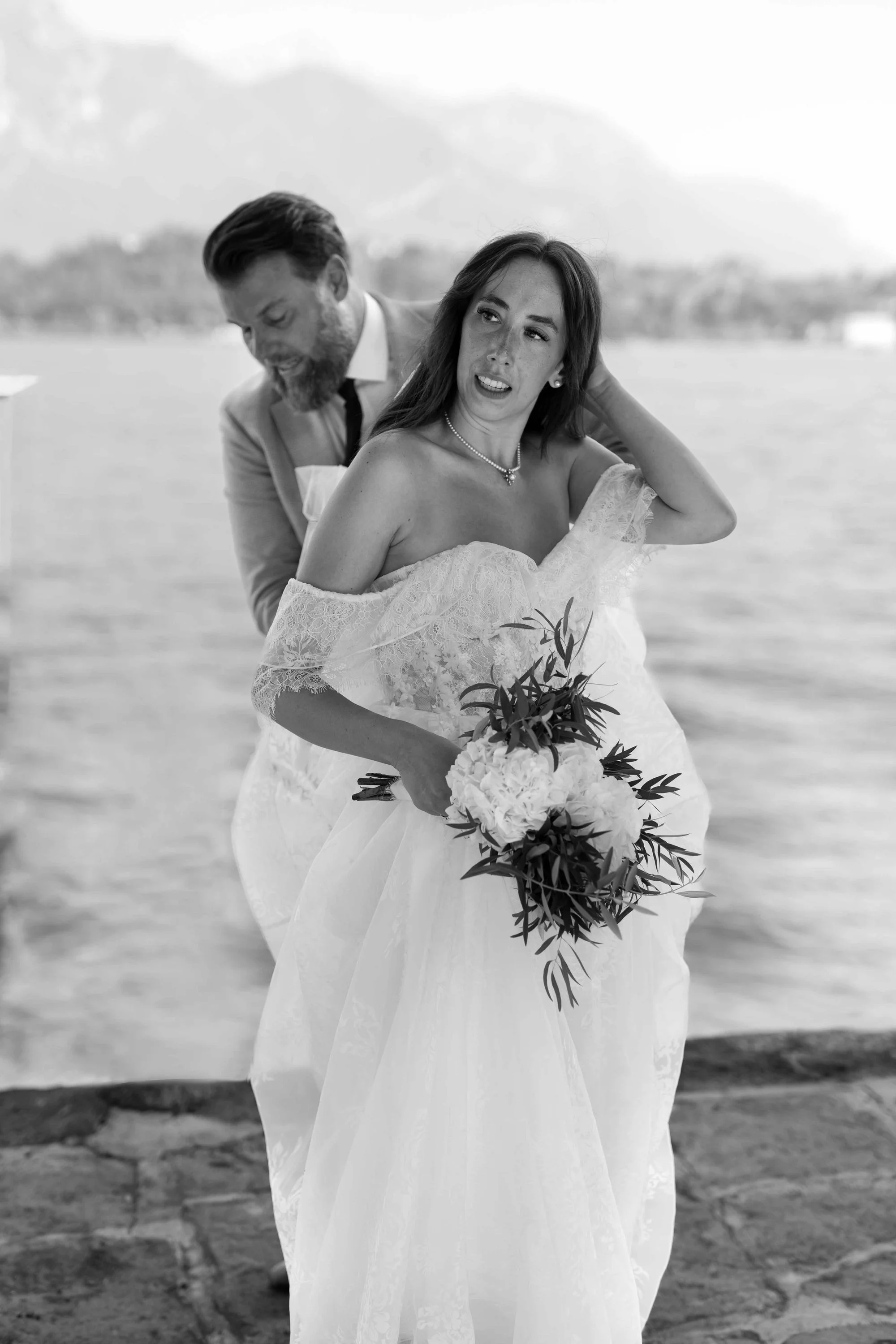 A black and white photo of a woman in a wedding dress holding a bouquet, standing by a body of water, with a man in a suit behind her, both outdoors.