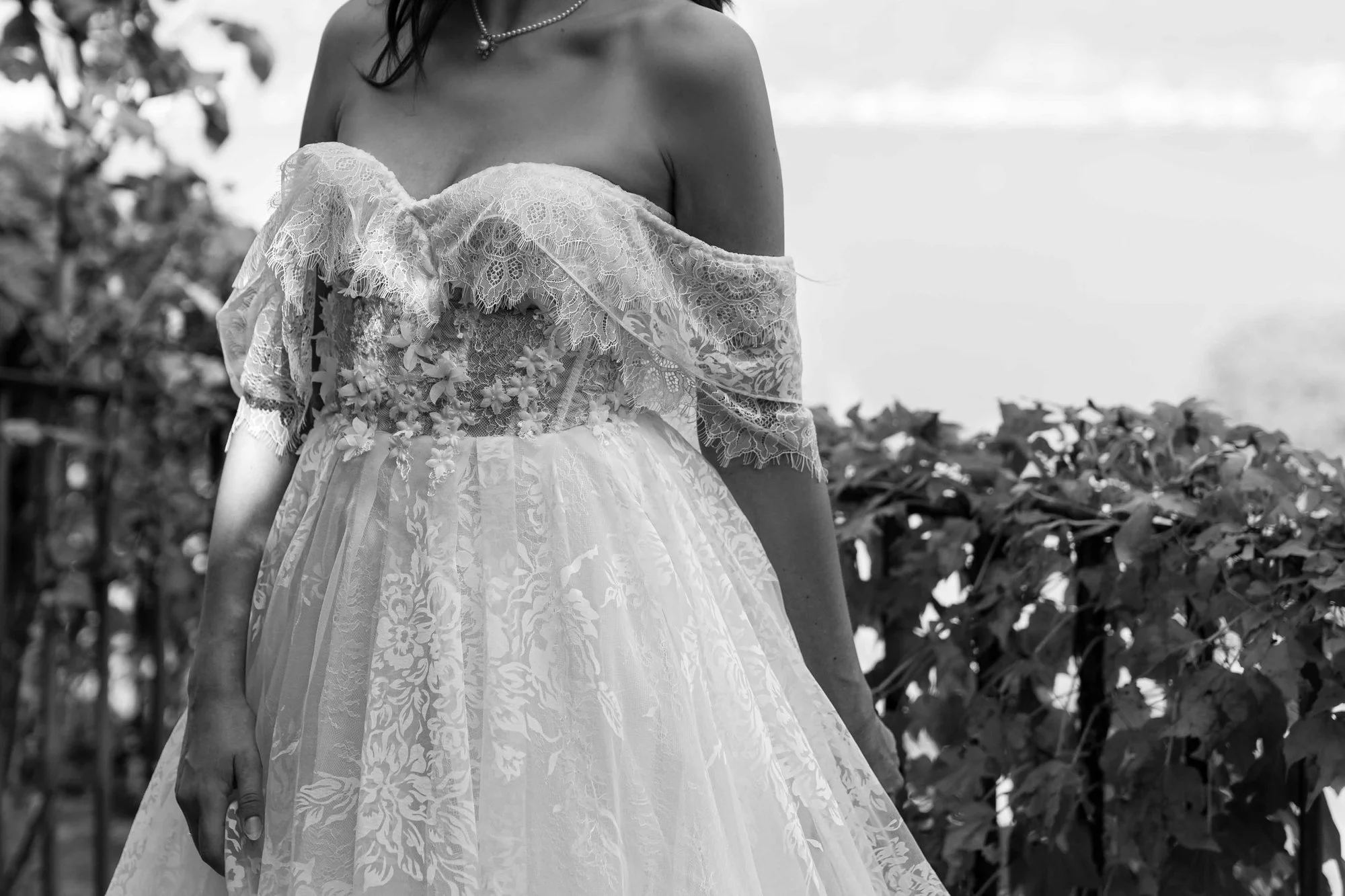 A woman wearing an off-the-shoulder lace and floral gown and a pearl necklace standing outdoors in front of leafy plants, captured in black and white.