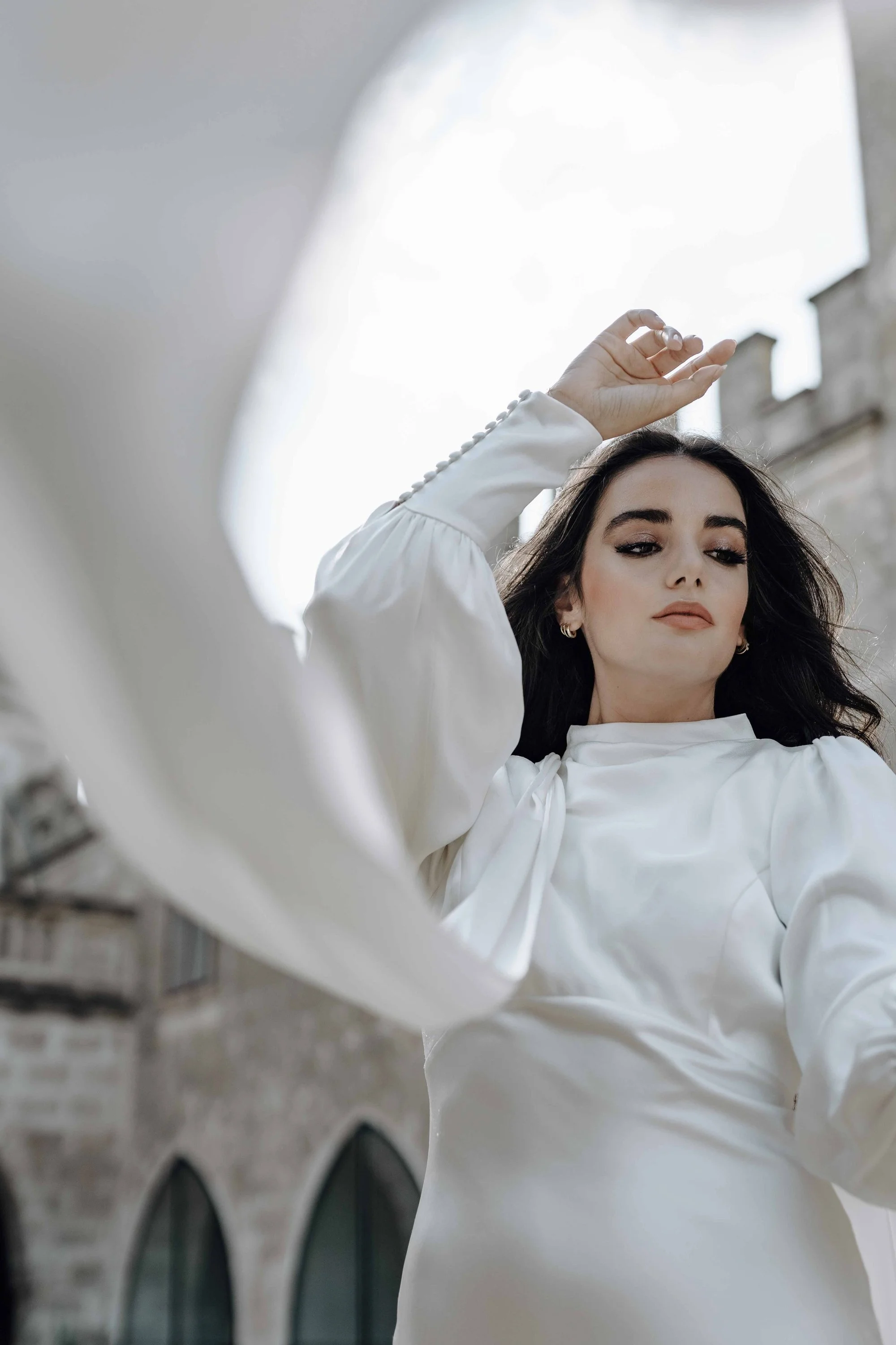A woman with dark hair and makeup, wearing a white long-sleeve blouse, looking down while raising her right hand, standing outdoors near a stone building with arched windows.