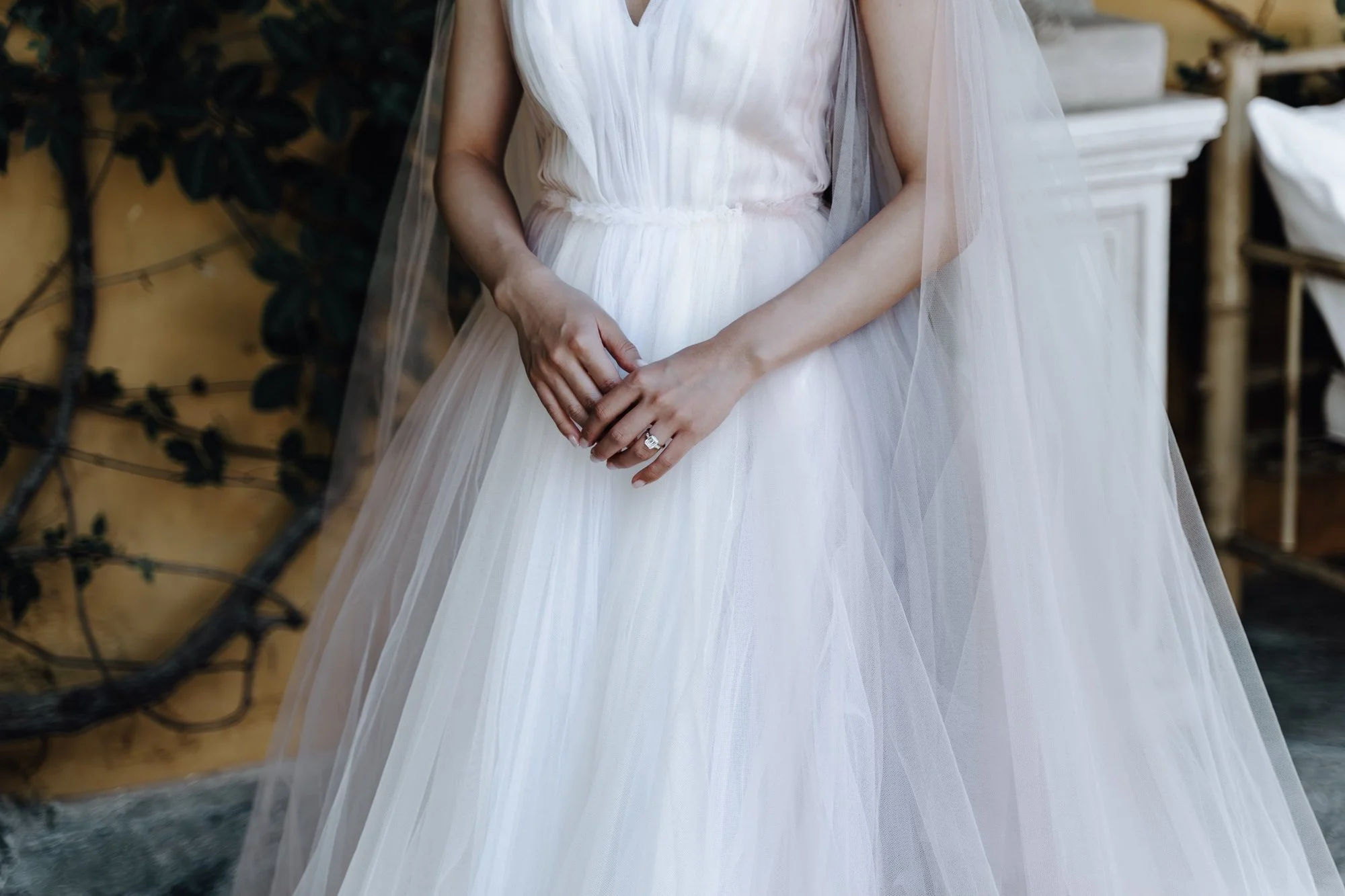 A woman in a white wedding dress with a long, flowing tulle skirt. She is standing outdoors with her hands clasped in front of her, showing a ring on her left hand. There are trees and outdoor furniture in the background.