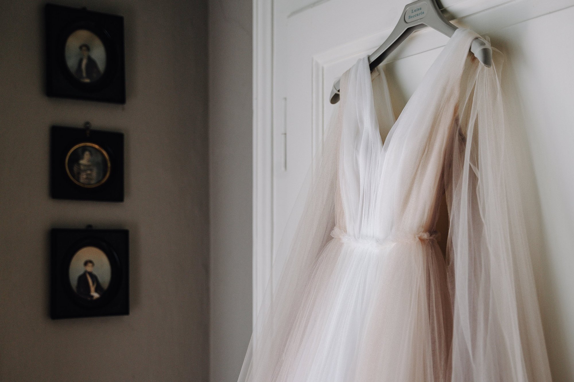 A white wedding dress hanging on a gray hanger labeled 'Lucia Beccari' on a door. In the background, there are framed photographs or artwork on the wall.