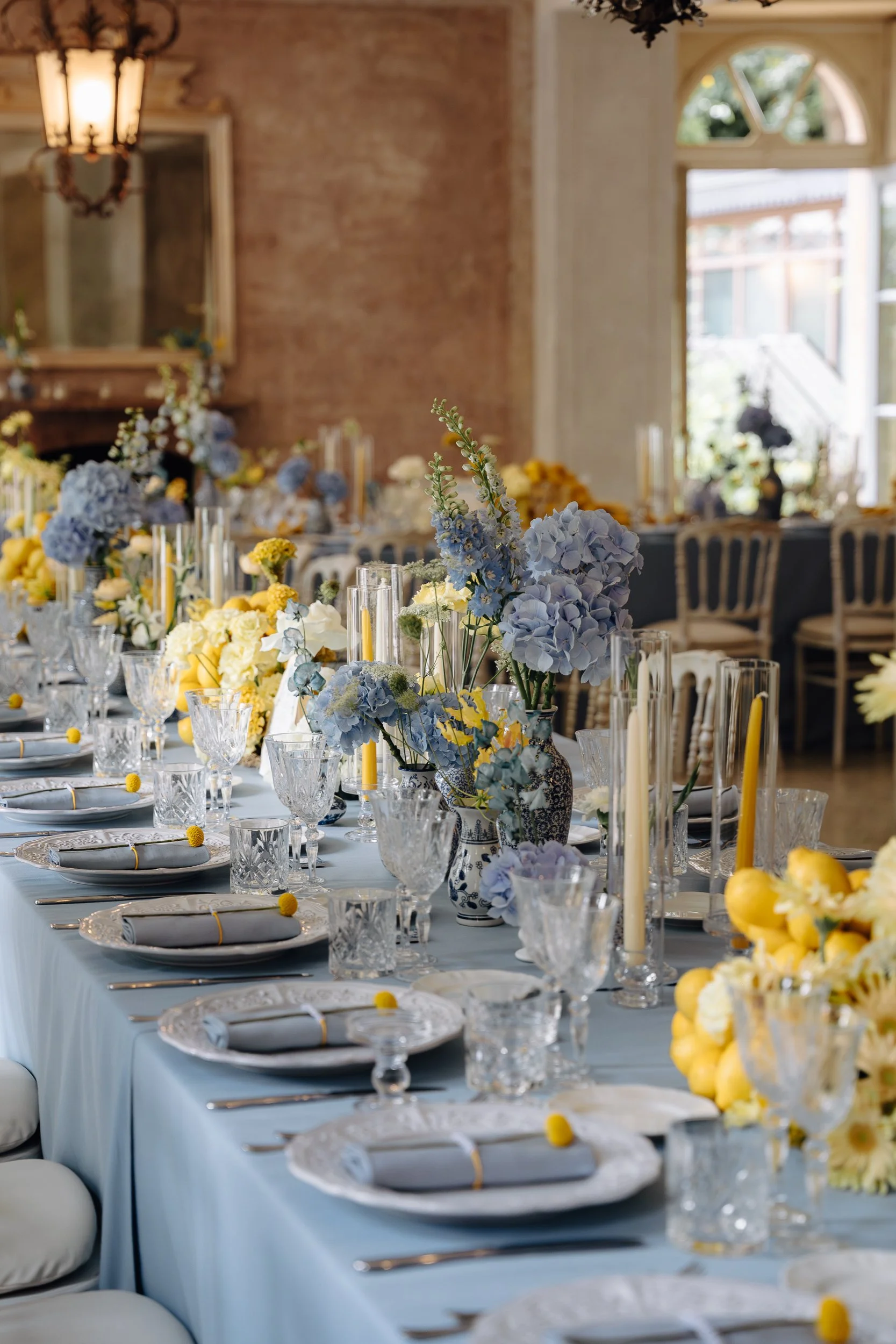 A beautifully decorated dining table with blue, white, and yellow flower arrangements, candles, and fine china in a bright room with large windows and ornate decor.