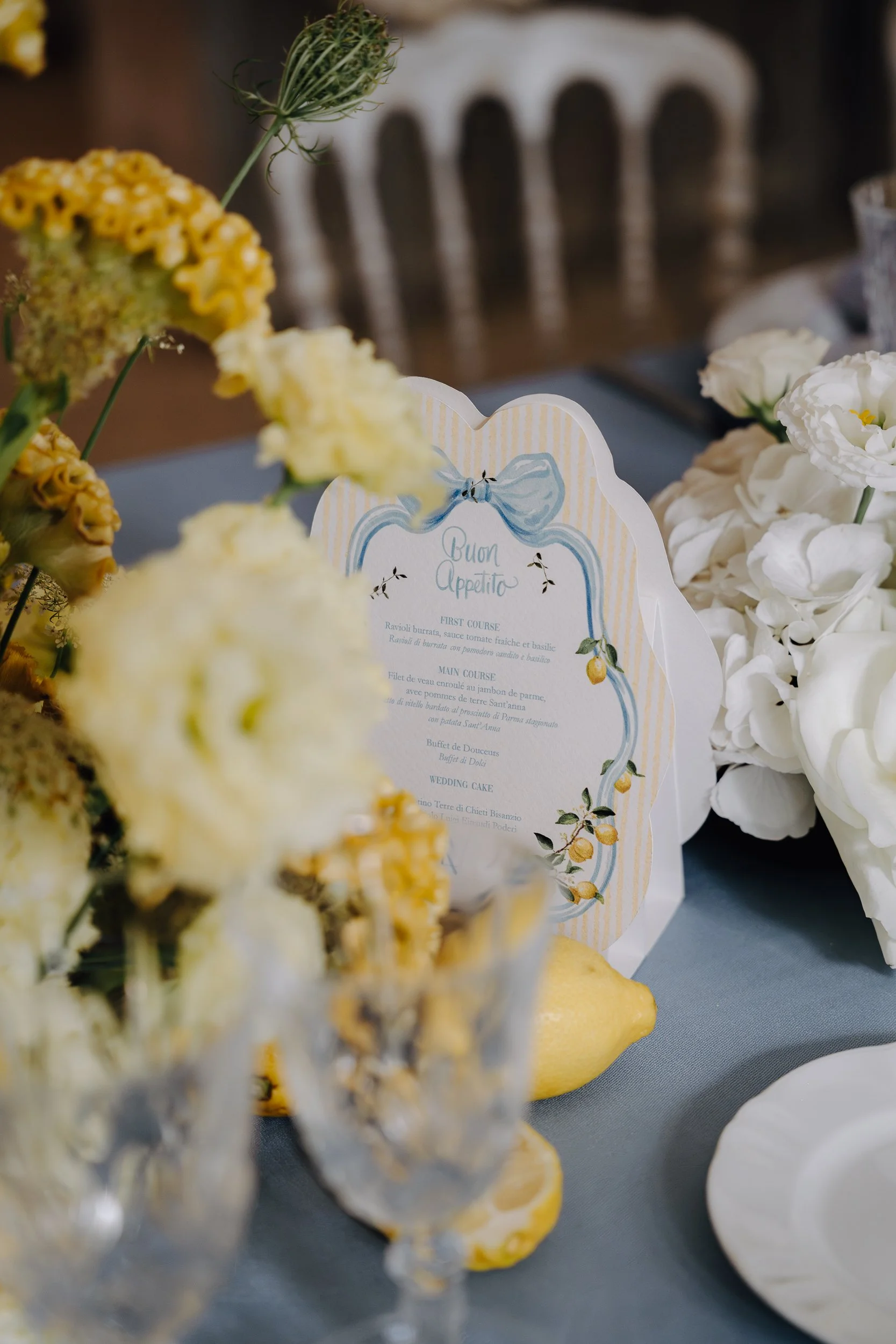 A wedding menu card surrounded by white and yellow flowers and a glass filled with yellow and white flowers.