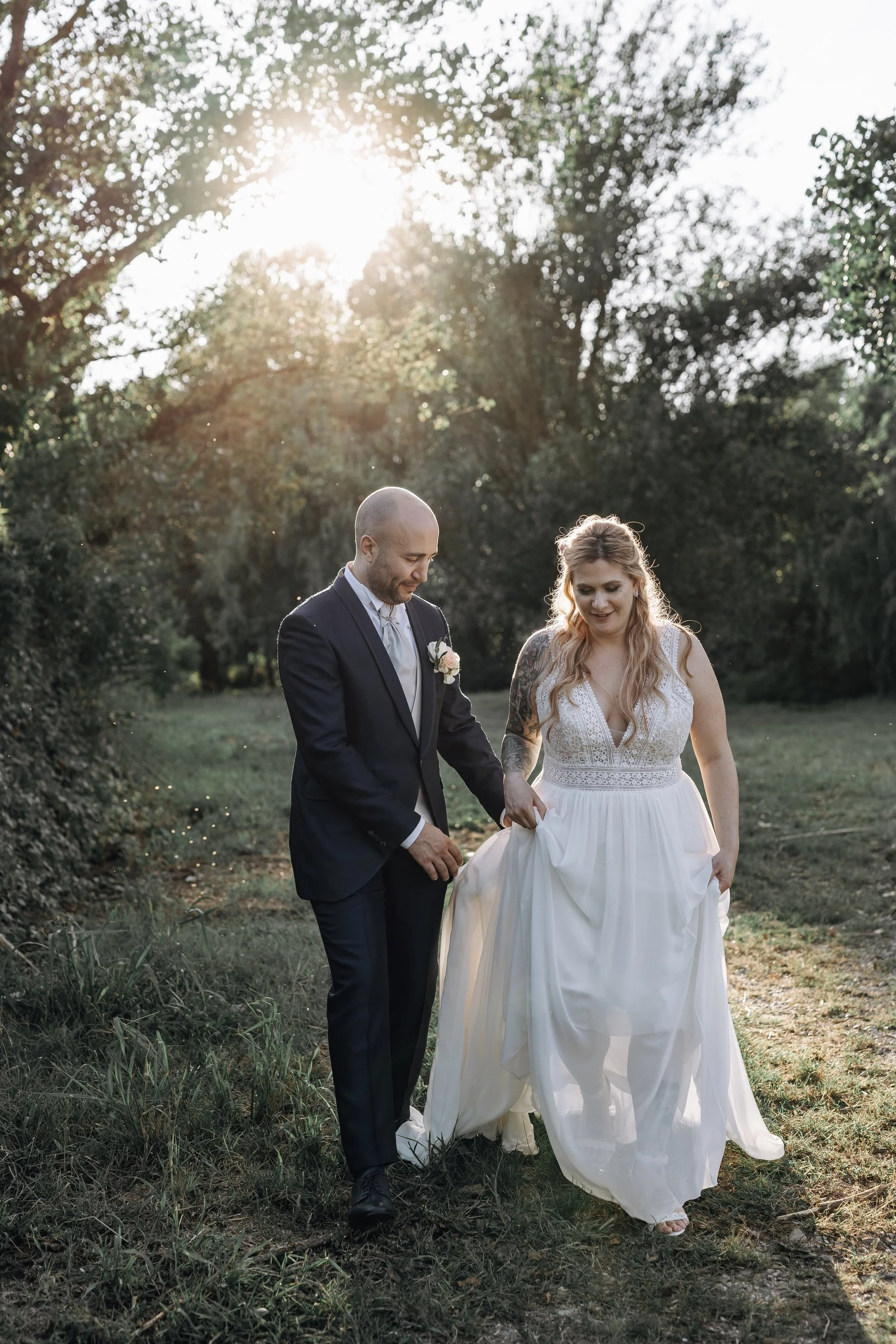 A bride and groom walking outdoors on a grassy area with trees in the background. The bride is holding up her white wedding dress while the groom is holding her hand. The sun is shining through the trees.