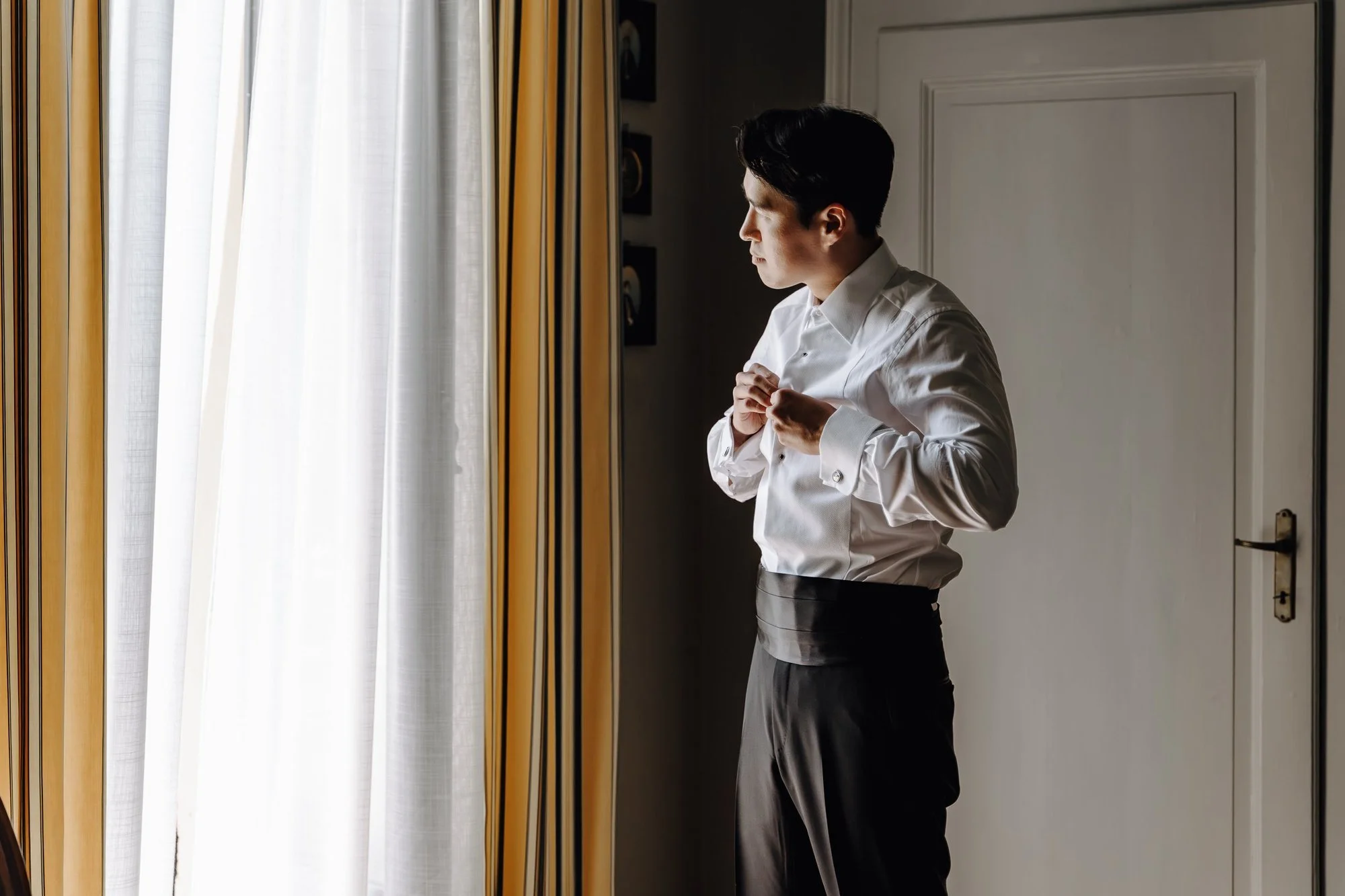 A man adjusting his white dress shirt in front of a mirror near a window with yellow and white curtains, in a room with white walls.