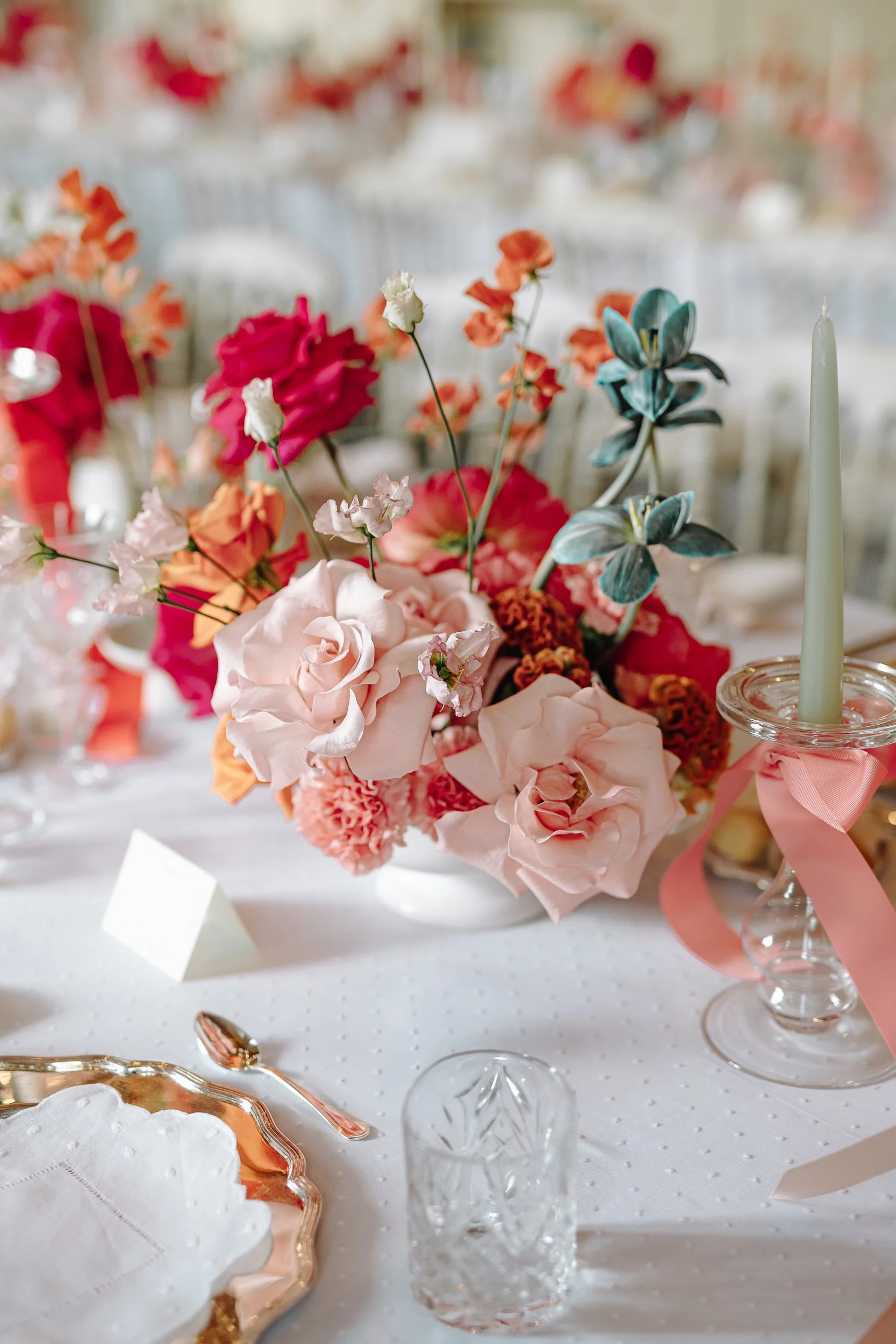 Elegant table setting with a centerpiece of pink, orange, and blue flowers in glass vases. Gold and pink accents on plates and cutlery, white tablecloth with subtle patterns. Pink ribbon decoration and a white candle.