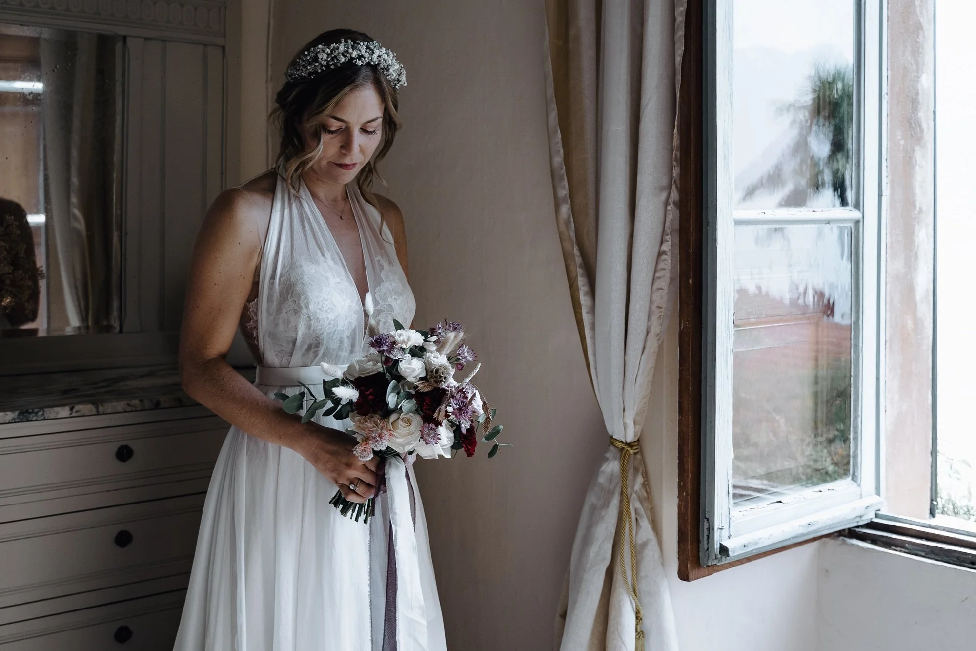 A bride standing indoors near a large window, holding a bouquet of flowers, wearing a white dress and a floral headband, with a neutral expression, in a room with beige walls and curtains.