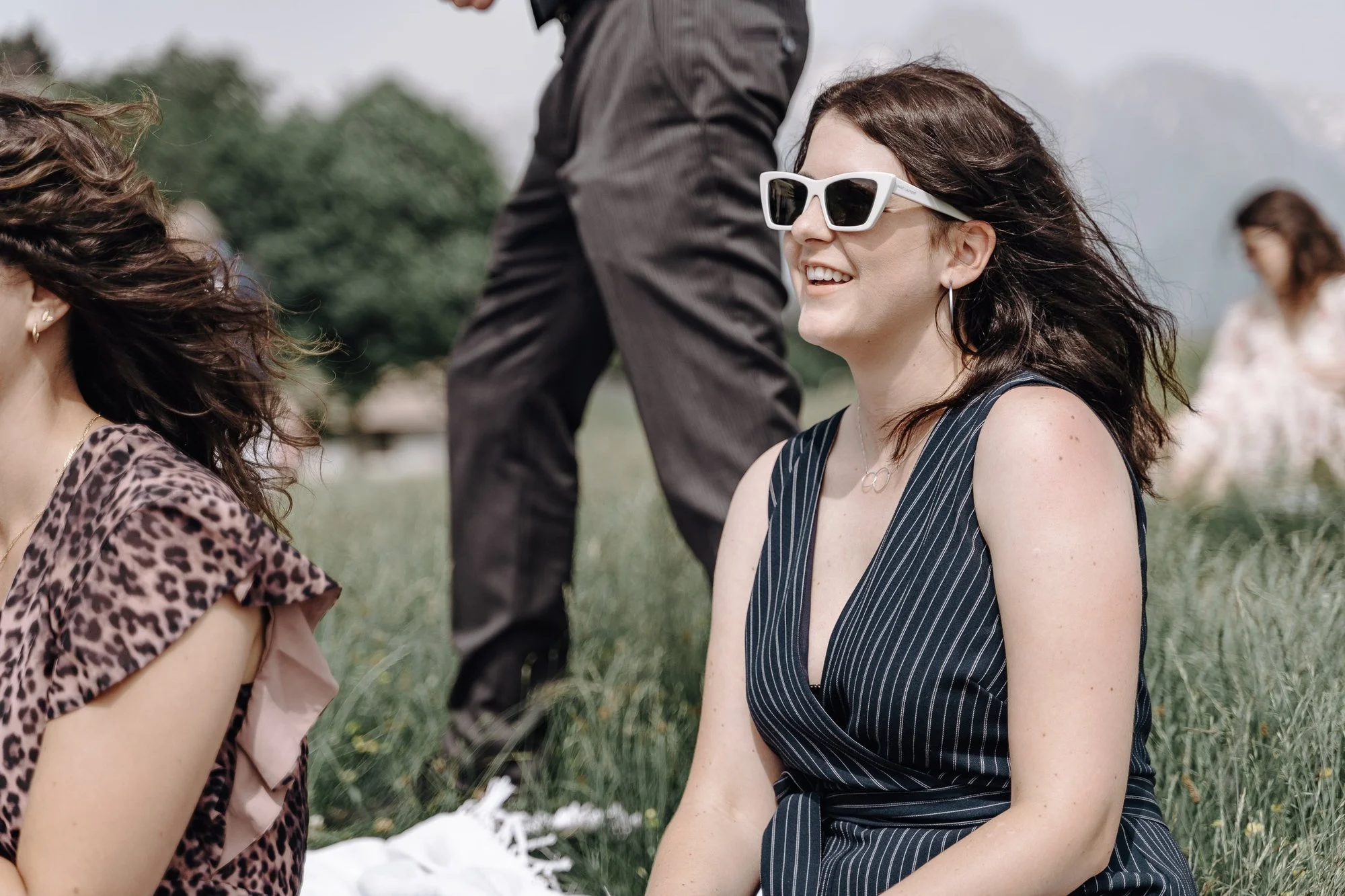 A woman with dark wavy hair wearing white sunglasses and a black striped dress smiling, sitting on grassy ground at an outdoor event with other people and trees in the background.