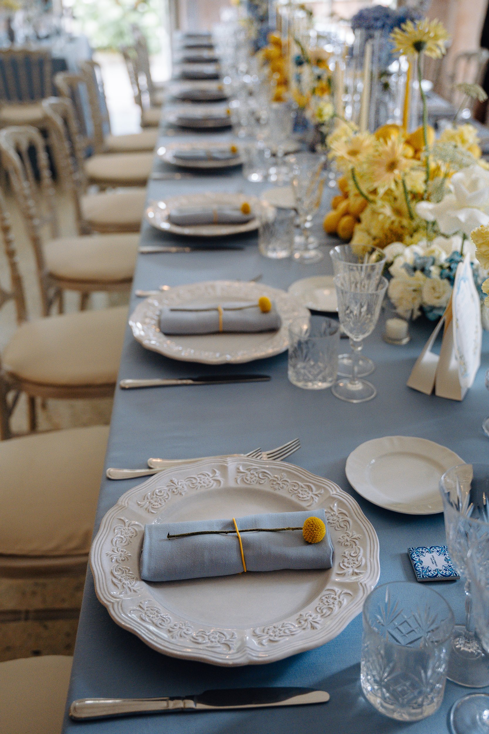 A long dining table set for a formal event with floral centerpieces, white plates with ornate edges, gray napkins with small yellow pom-pom, silver utensils, and clear glasses, with beige chairs surrounding it.