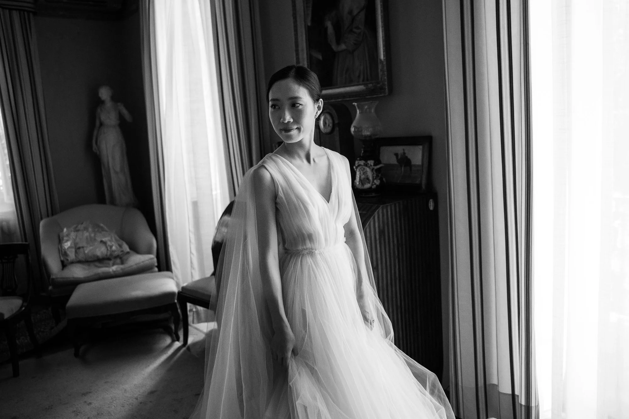 A woman in an elegant, flowing dress stands in a room with vintage decor, gazing to the side. The room has a large window with light curtains, a sofa with a piece of cloth on it, and artwork on the walls.
