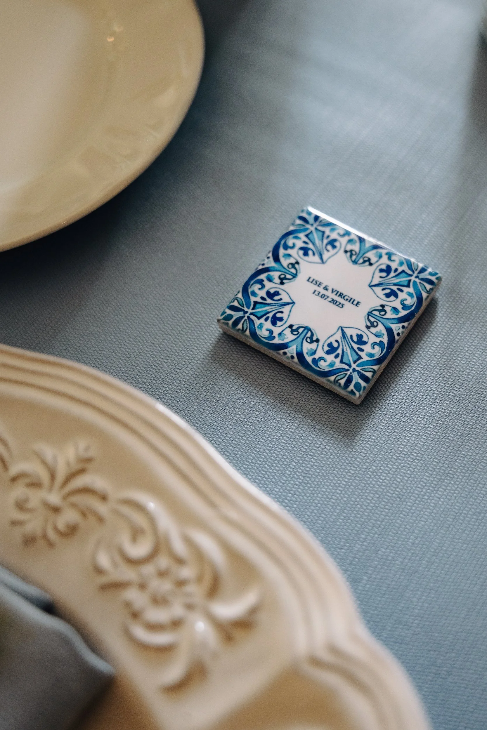 Close-up of a decorative ceramic box with blue and white design and the words "Use & Virgile 13/07/2025" on the lid, placed on a textured gray surface, with a beige ornate object in the foreground.