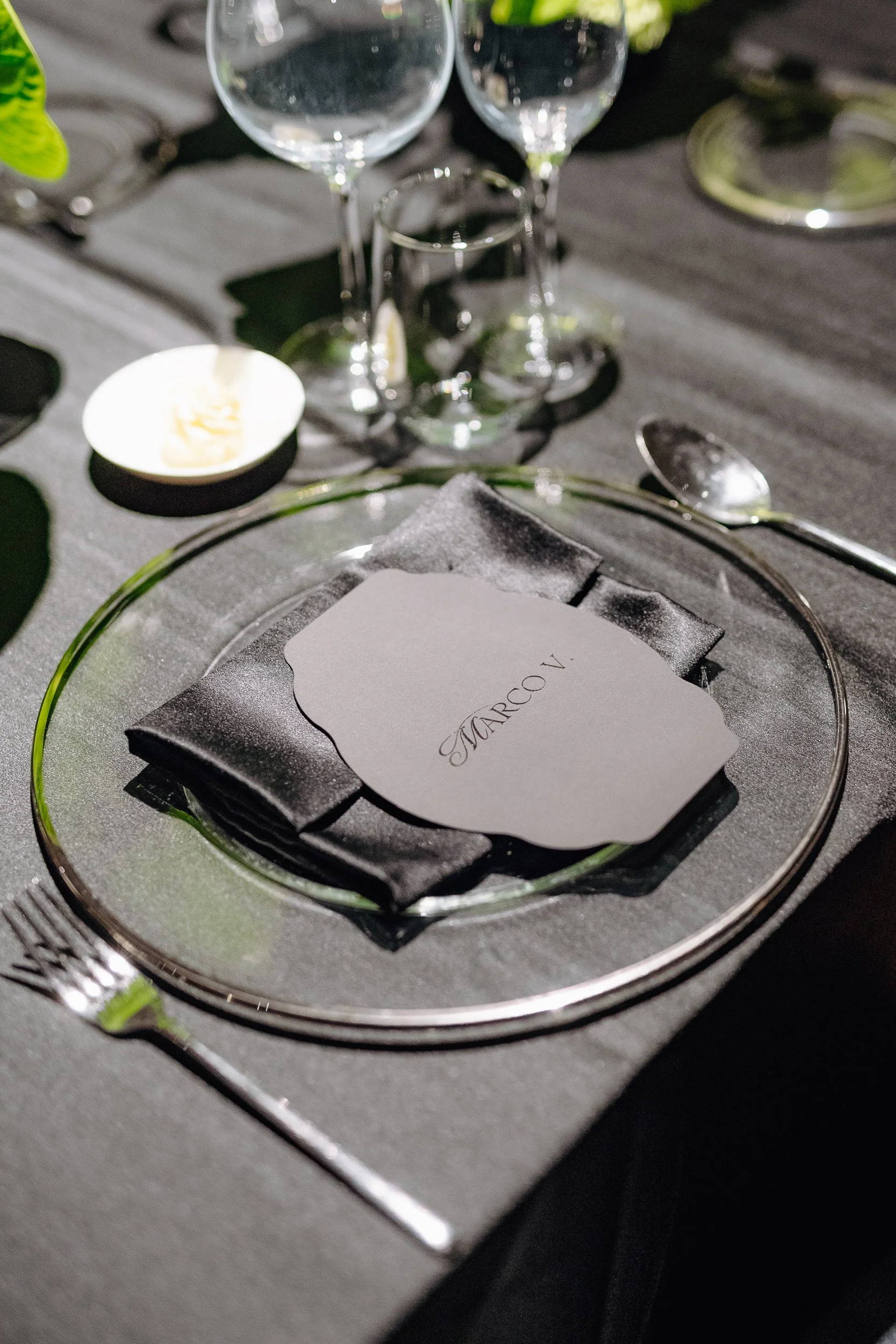 Close-up of a dinner table setting with a black napkin, a silver spoon, a glass plate, and a black box with the word "MARCO" written on it. There are two empty wine glasses and a small lit candle in the background.