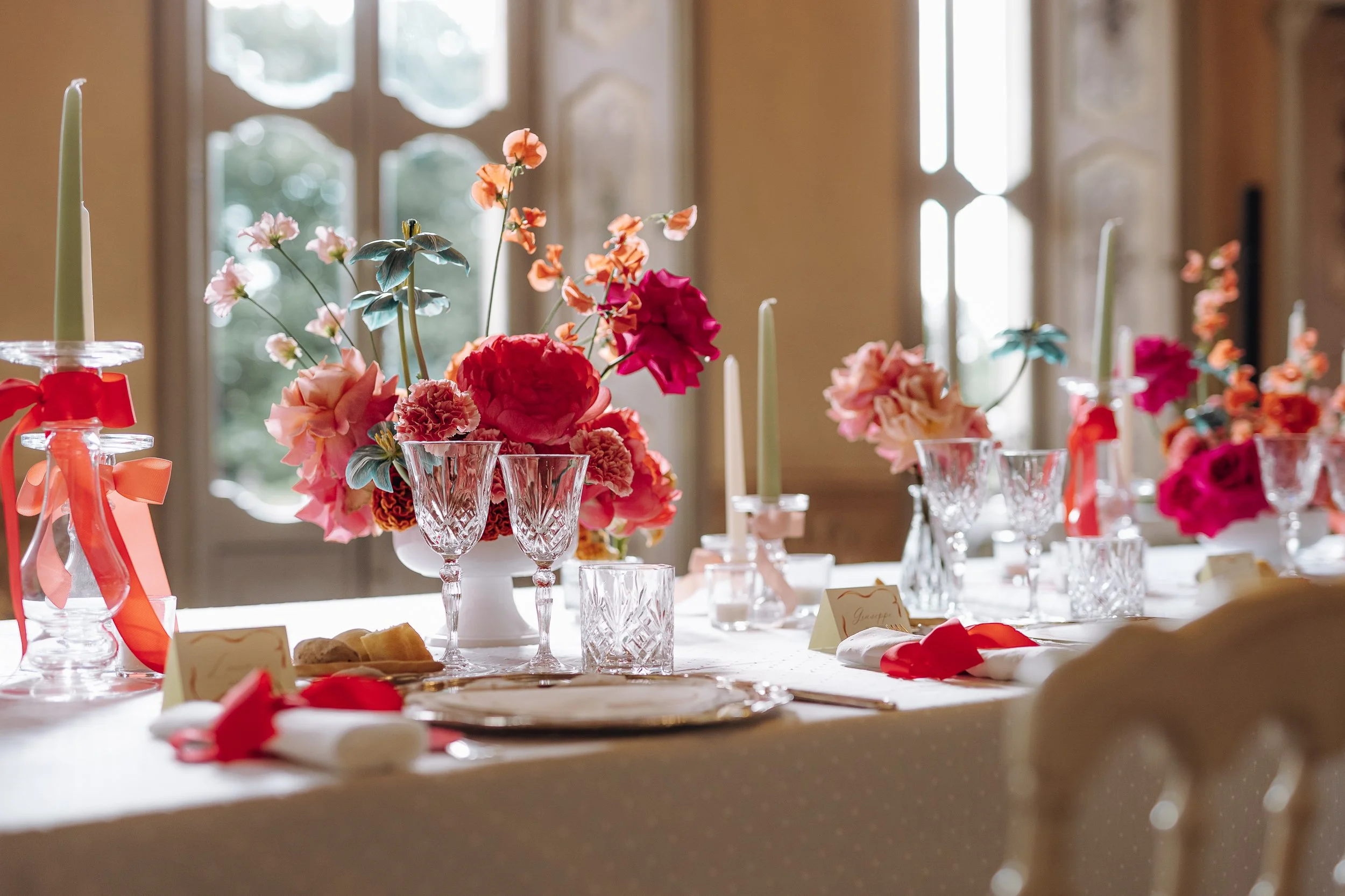 A wedding or special event table decorated with pink and red floral arrangements in glass vases, with white candles and place cards, set near large windows that let in natural light.