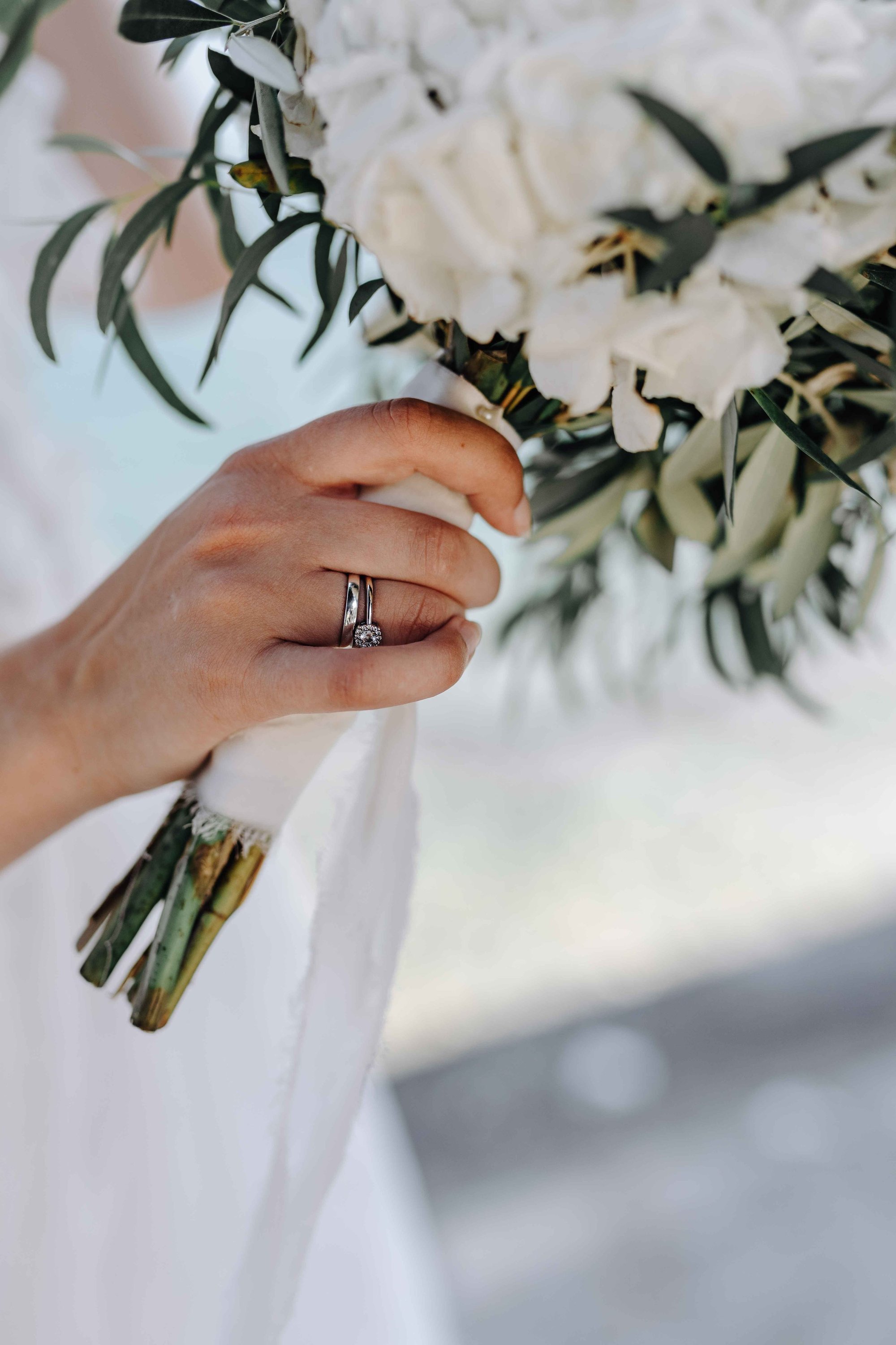 A hand with a wedding ring holding a bouquet of white flowers and dark green leaves.