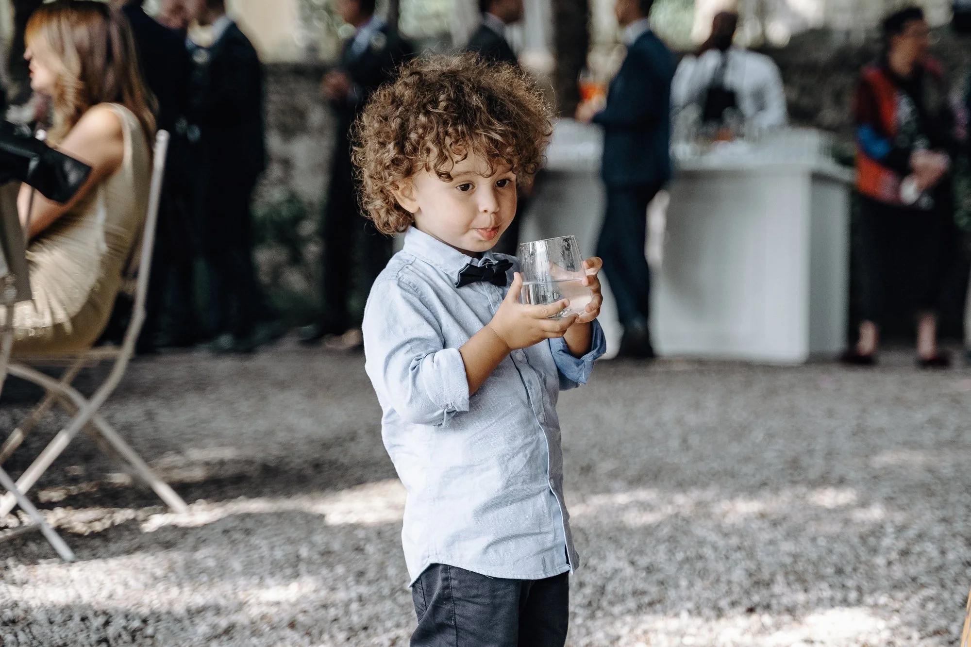 A young boy with curly hair holding a glass of water at an outdoor event with other people and a food station in the background.