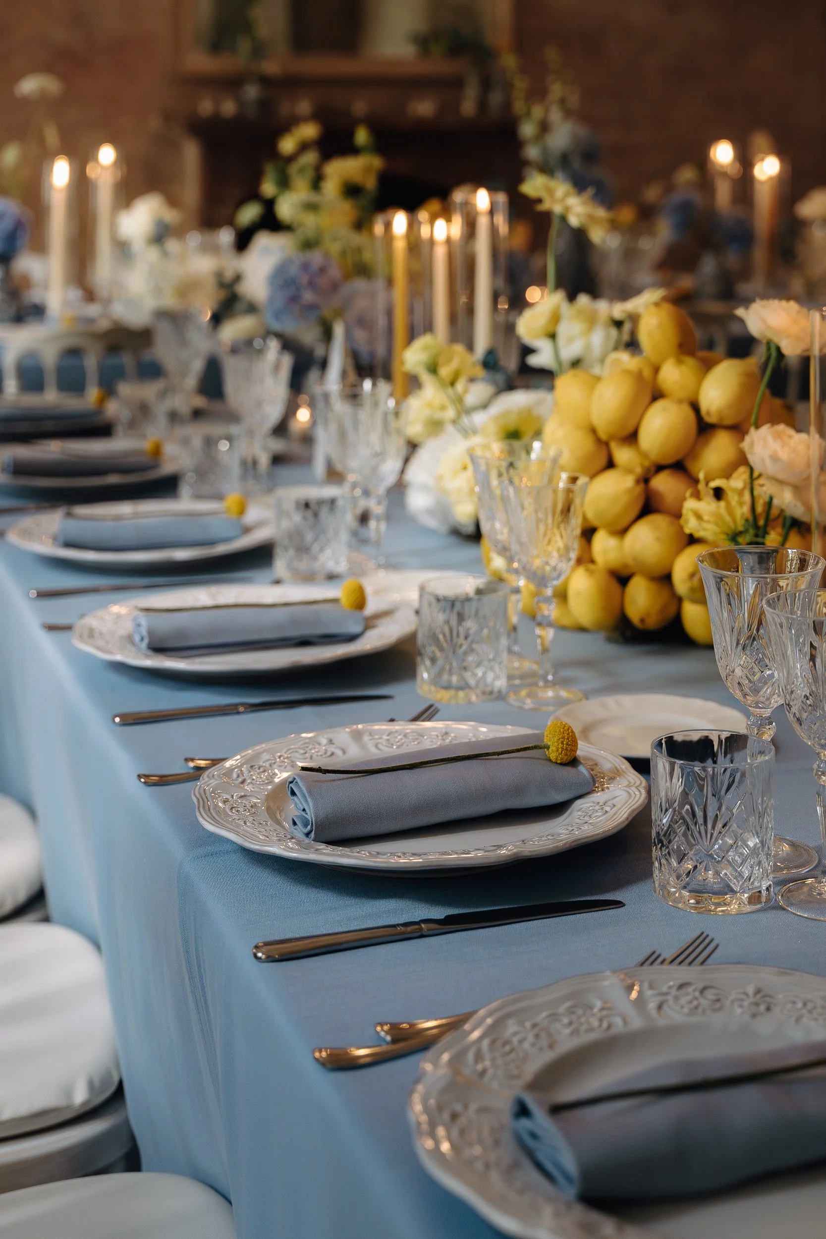 Elegant banquet table set with white and blue tablecloth, white plates with ornate rims, light blue napkins, silver cutlery, and intricate glassware. The table is decorated with a large floral centerpiece featuring yellow lemons, white and light purp