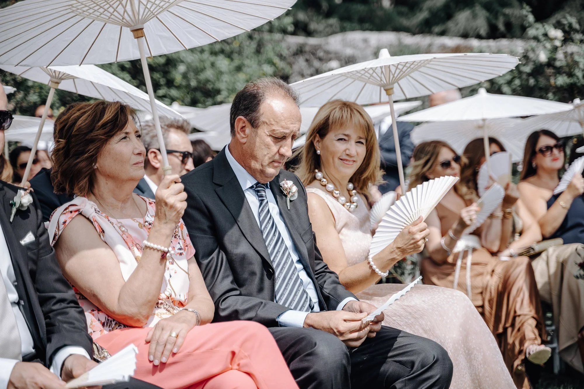 A group of people seated outdoors during a formal event, holding white parasols, with some reading or holding papers, dressed in formal attire, with a blurred natural background.