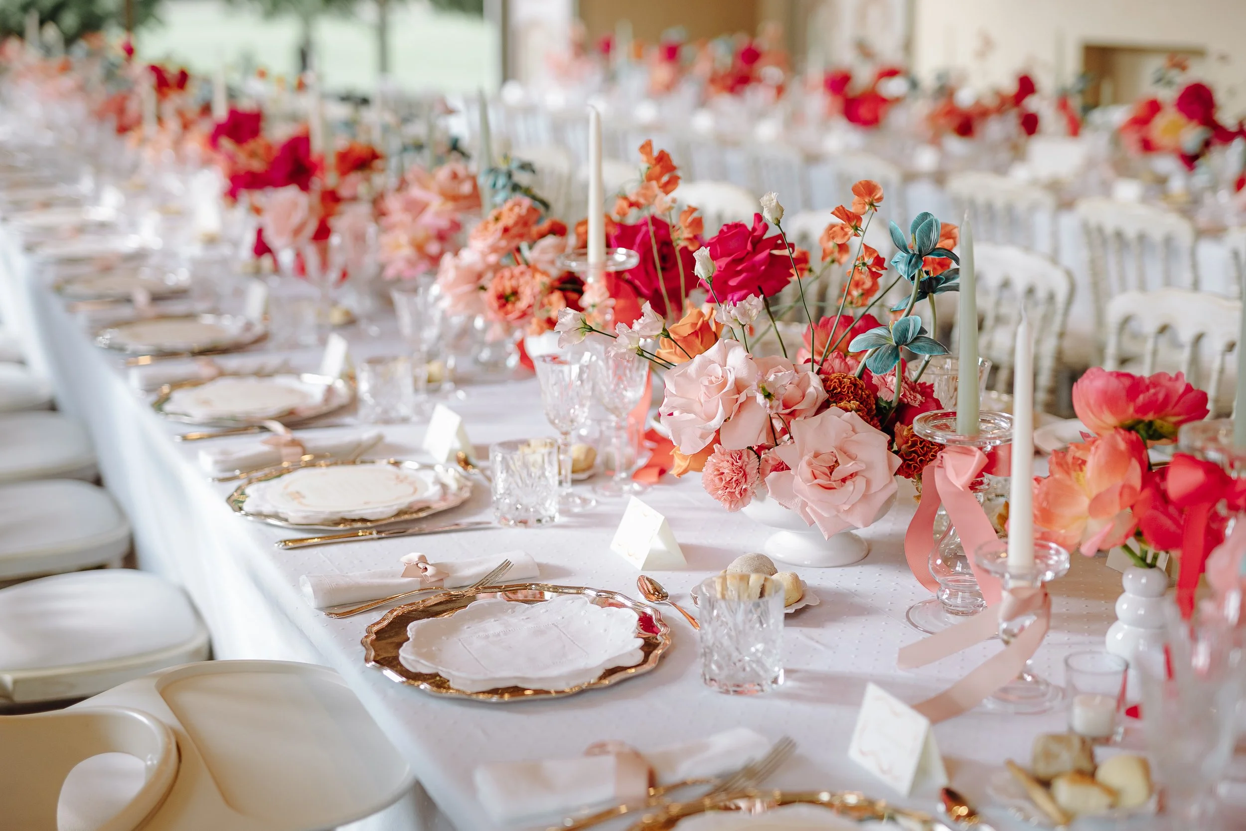 A long banquet table decorated with pink, coral, and blue flowers, white candles, and fine china for a celebration or wedding reception.