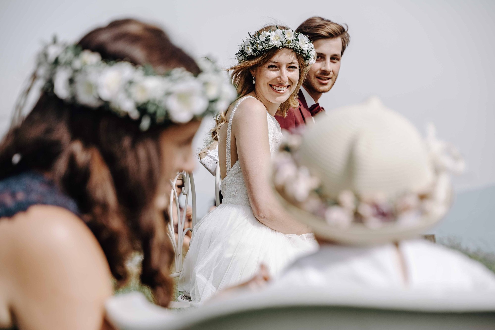 A smiling bride with a floral crown sitting among friends at her wedding, with friends partly visible in the foreground.