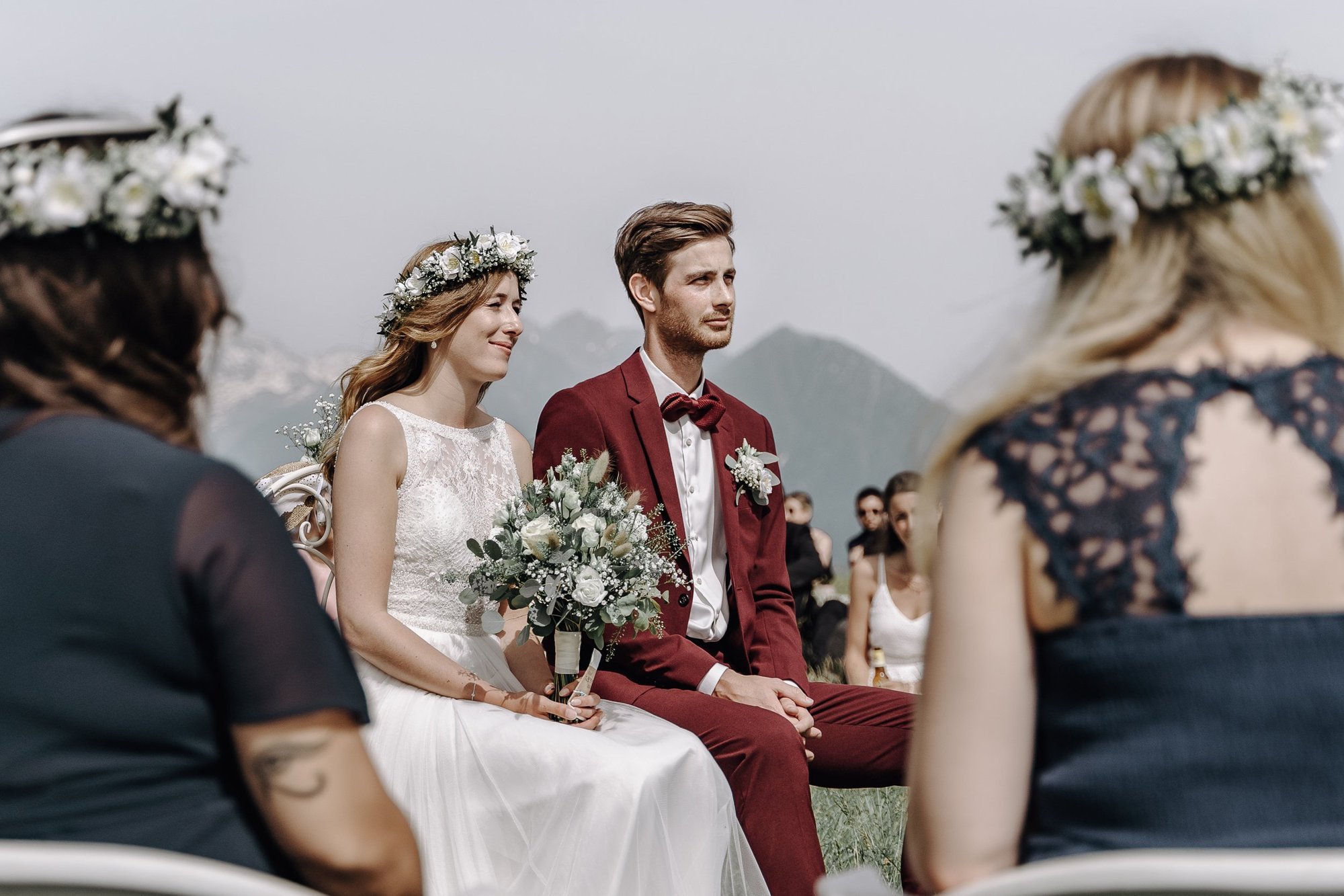 A bride and groom sit side by side during an outdoor wedding ceremony, surrounded by guests, with mountains in the background. The bride wears a white lace dress and a floral crown, holding a bouquet of white flowers. The groom wears a burgundy suit 