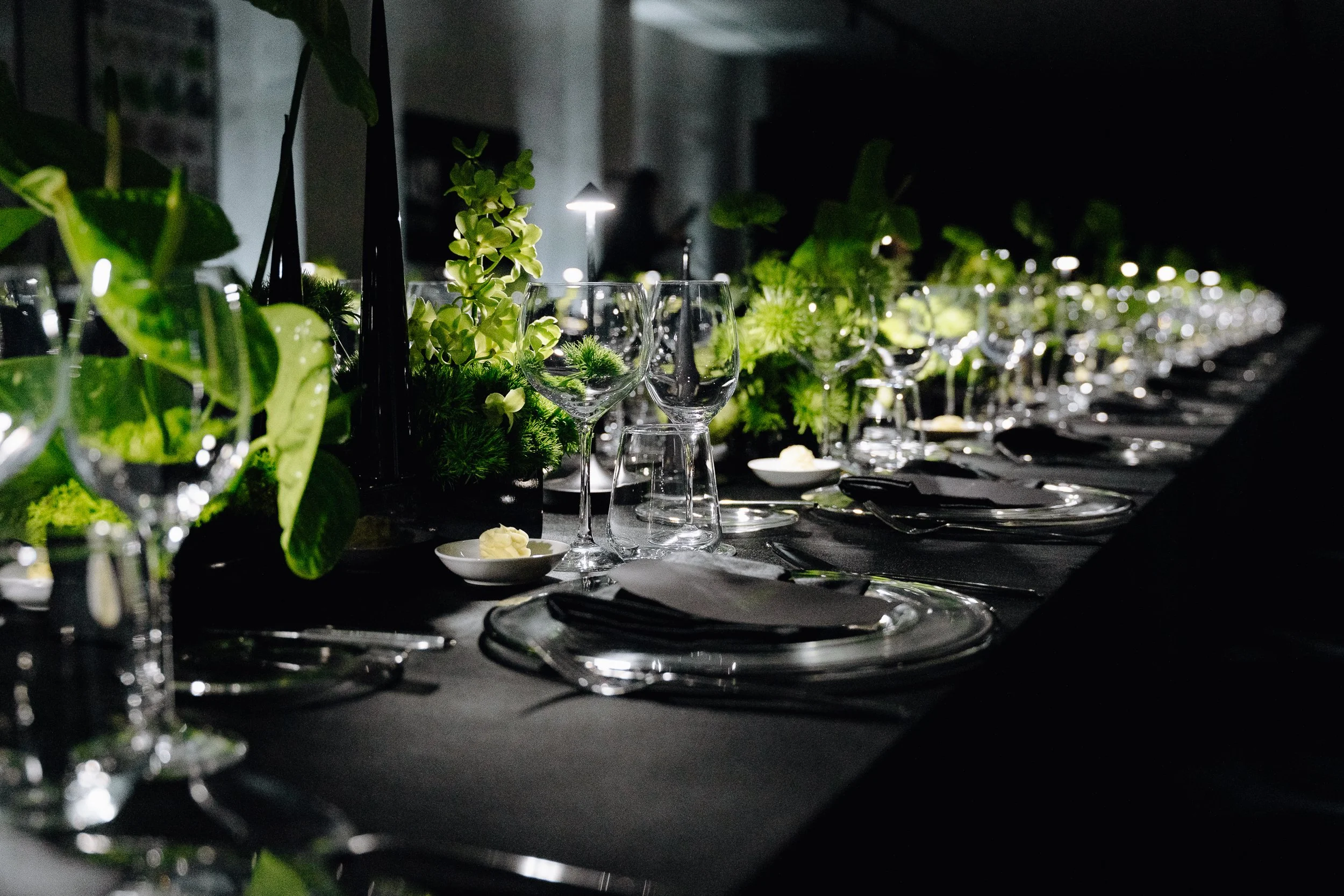 A long banquet table decorated with lush green plants, glassware, and small plates of butter, set in a dimly lit venue for an elegant event.