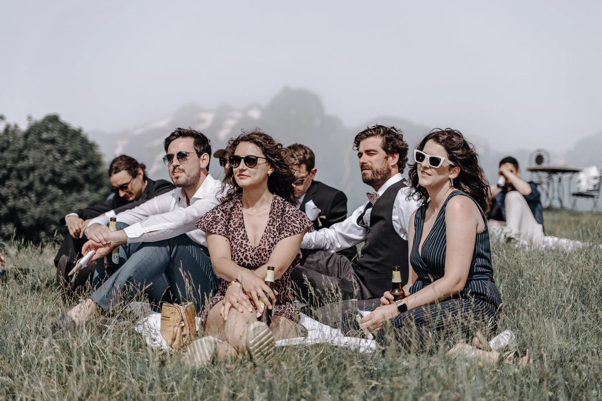 A group of people sitting on the grass outdoors, with a mountain in the background. They are dressed semi-formally, some wearing sunglasses, and appear to be enjoying a sunny day.
