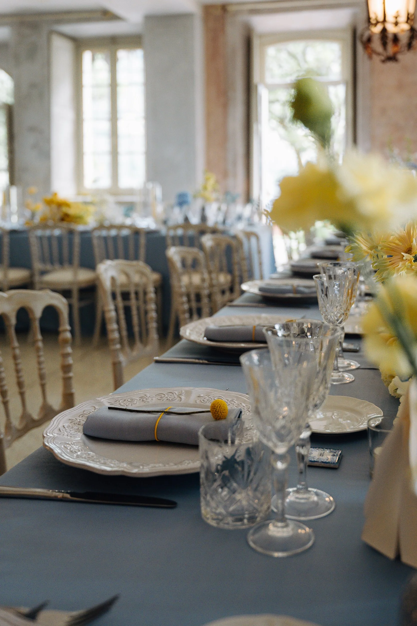 A long table set for a formal event with ornate white plates, gray napkins secured with yellow pom-pom napkin rings, and crystal glassware. The table is decorated with floral arrangements of yellow flowers. In the background, there are wooden chairs 