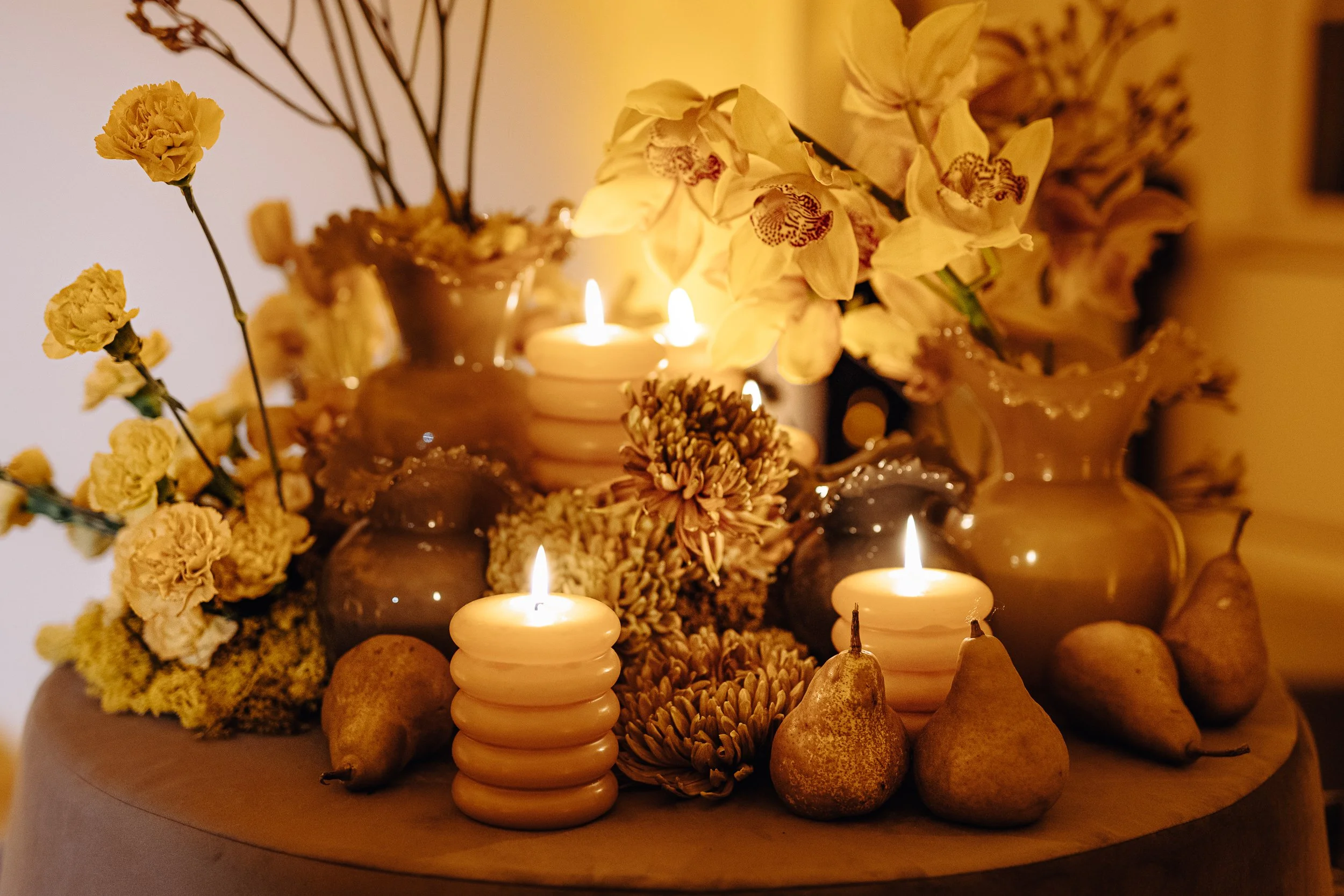 A floral arrangement featuring yellow flowers, including orchids, with lit candles, decorative vases, and pears on a round table, creating a warm and elegant ambiance.