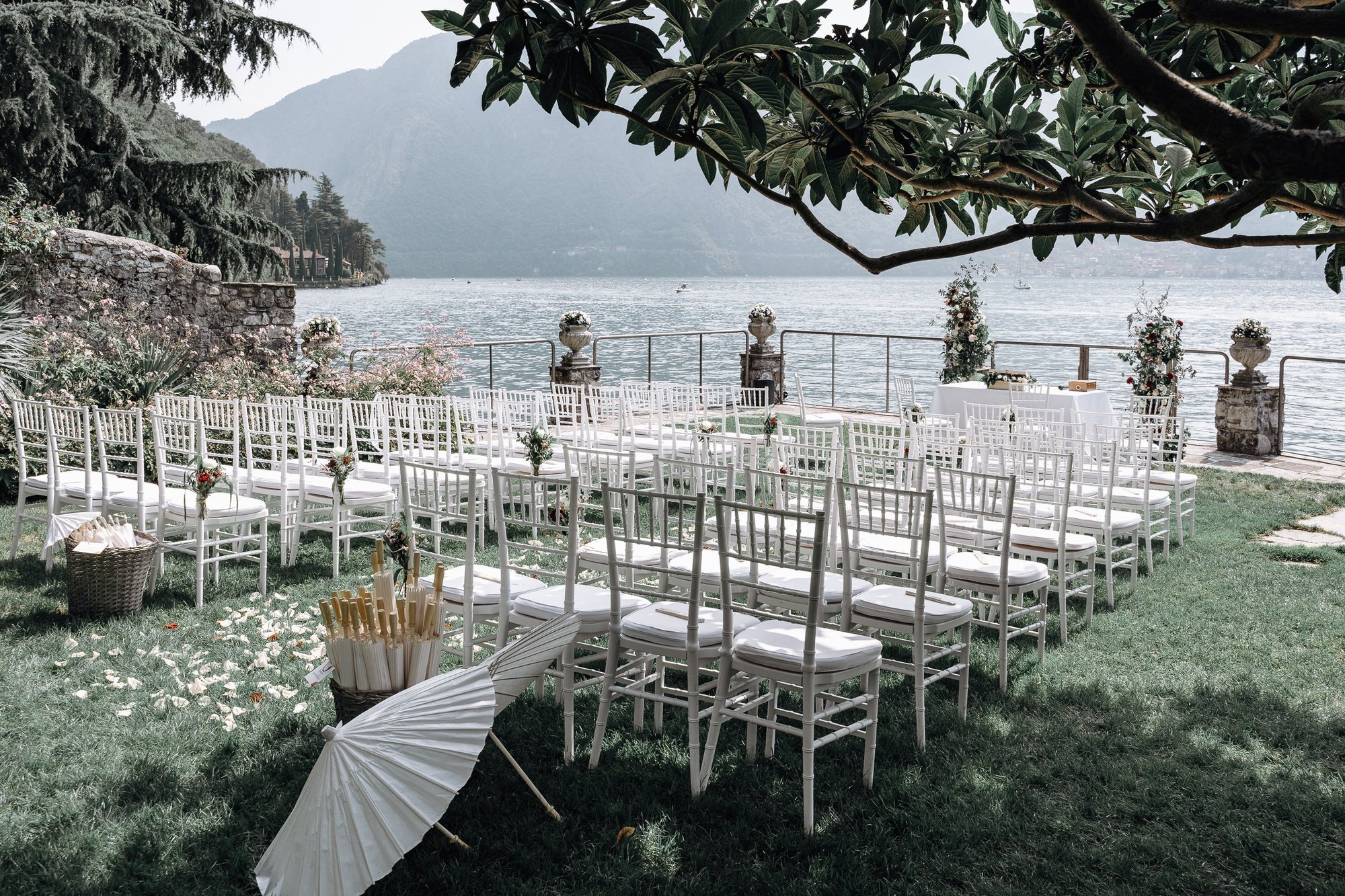 Outdoor lakefront wedding setup with white chairs arranged on green grass, floral decorations, and a parasol on the ground, overlooking a calm lake surrounded by mountains and trees.