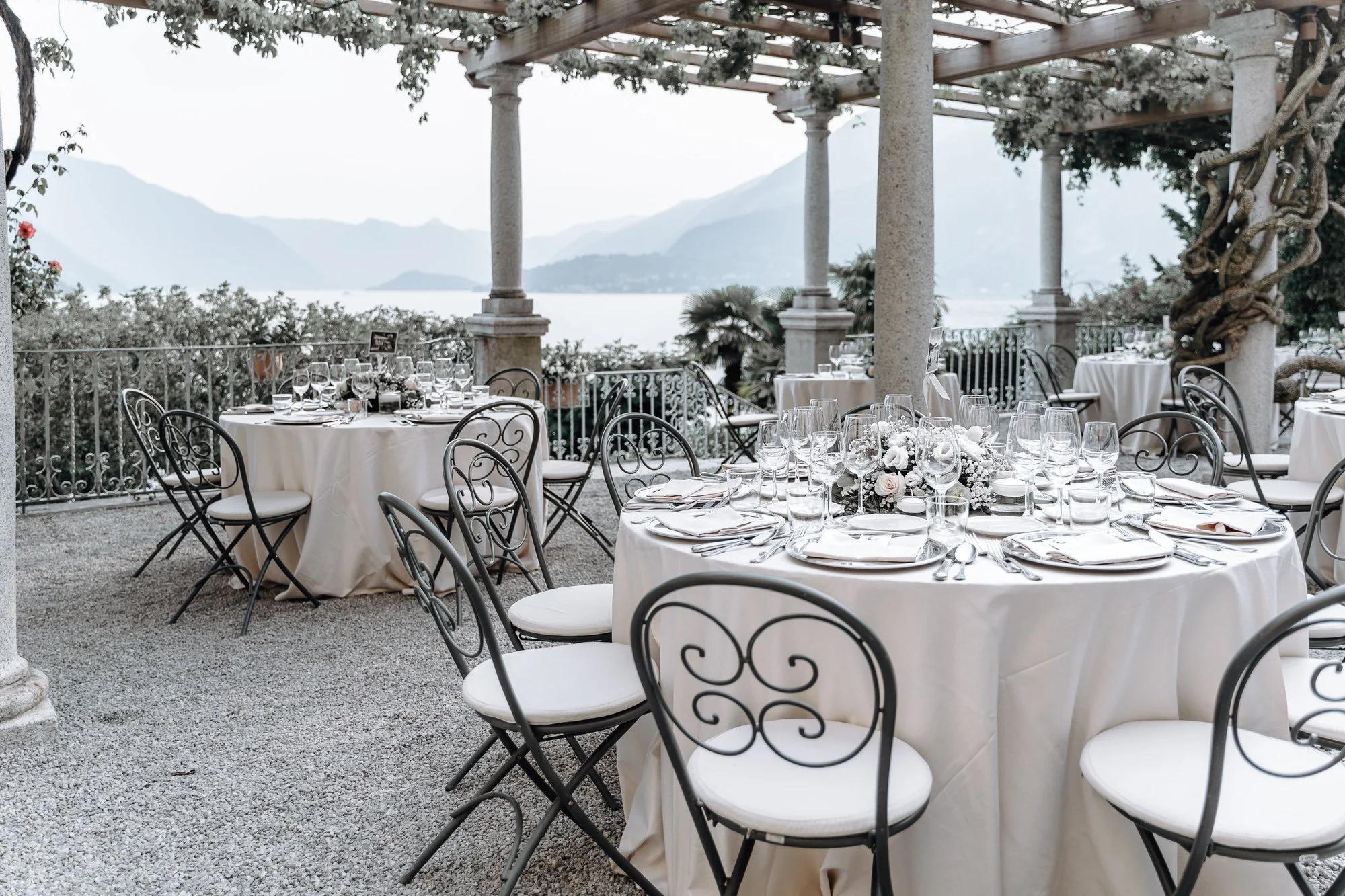 Elegant outdoor dining setup with round tables covered in white tablecloths, set with glassware and floral centerpieces, under a pergola overlooking a scenic body of water and mountains.