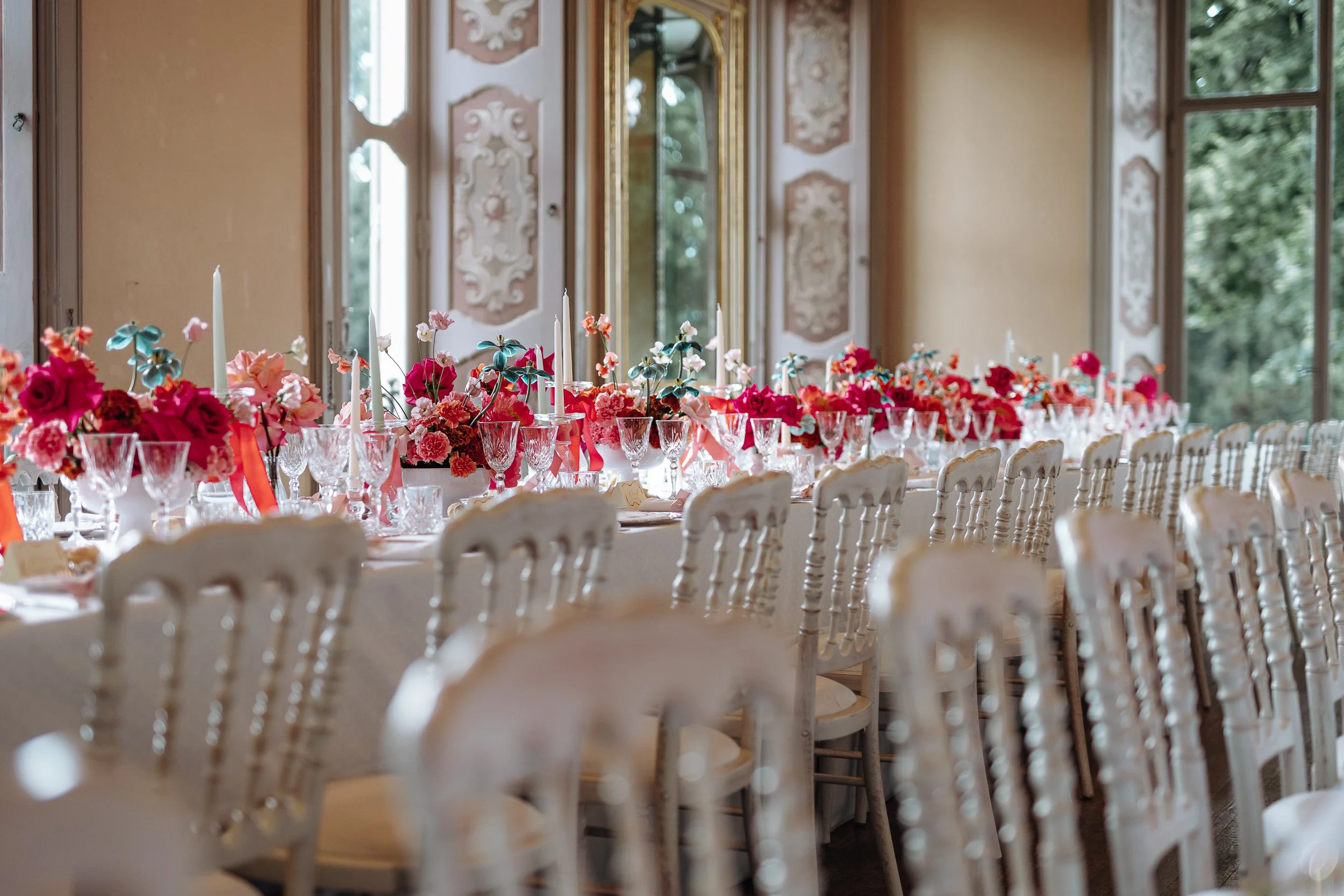 A long banquet table decorated with pink and red flowers and tall white candles, set up for a formal event in a bright room with large windows and ornate wall decorations.