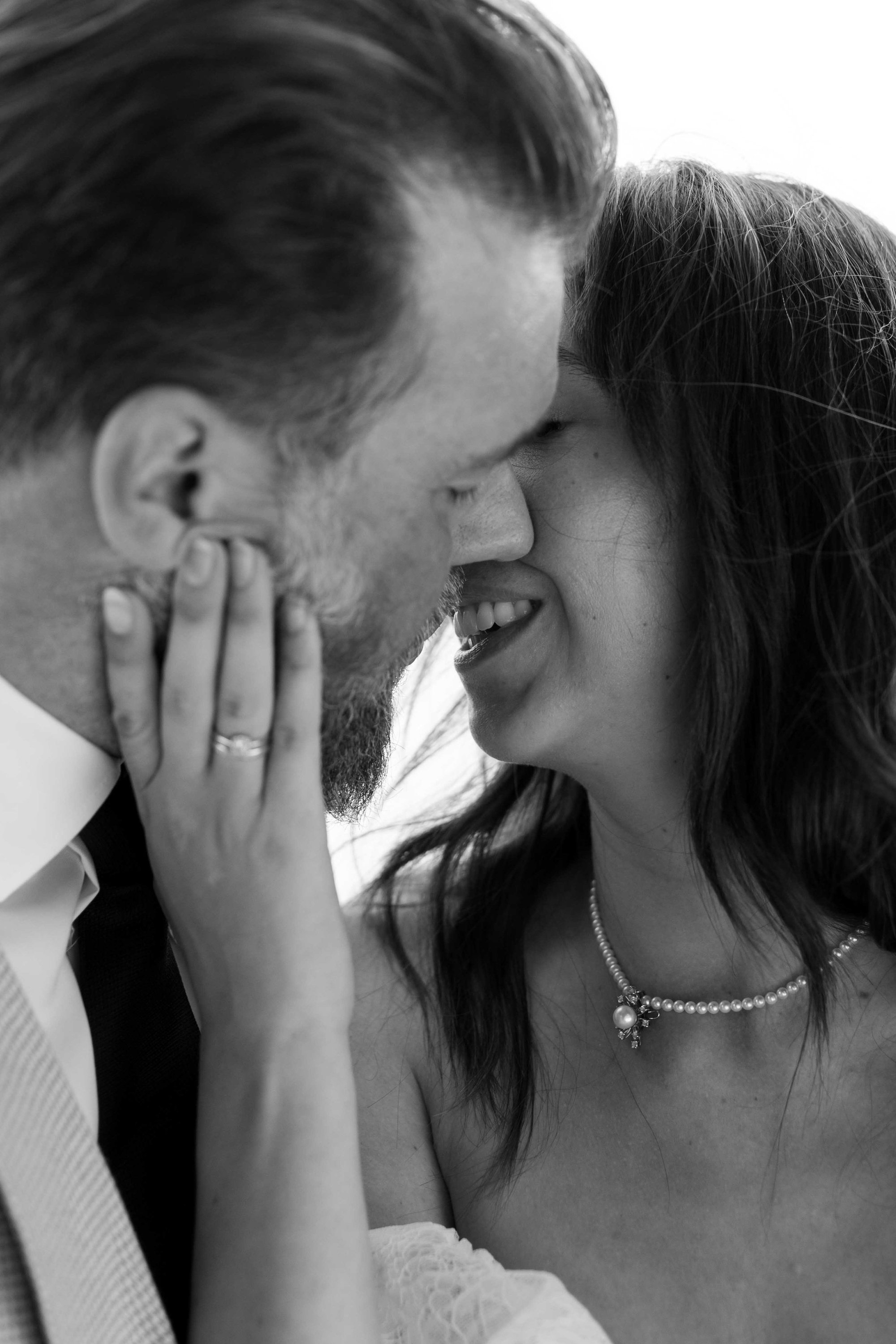 A black and white photo of a couple sharing an intimate moment, touching foreheads with closed eyes, both smiling gently. The woman has long hair, wears a pearl necklace, and the man has a beard and mustache. The woman's hand gently touches the man's