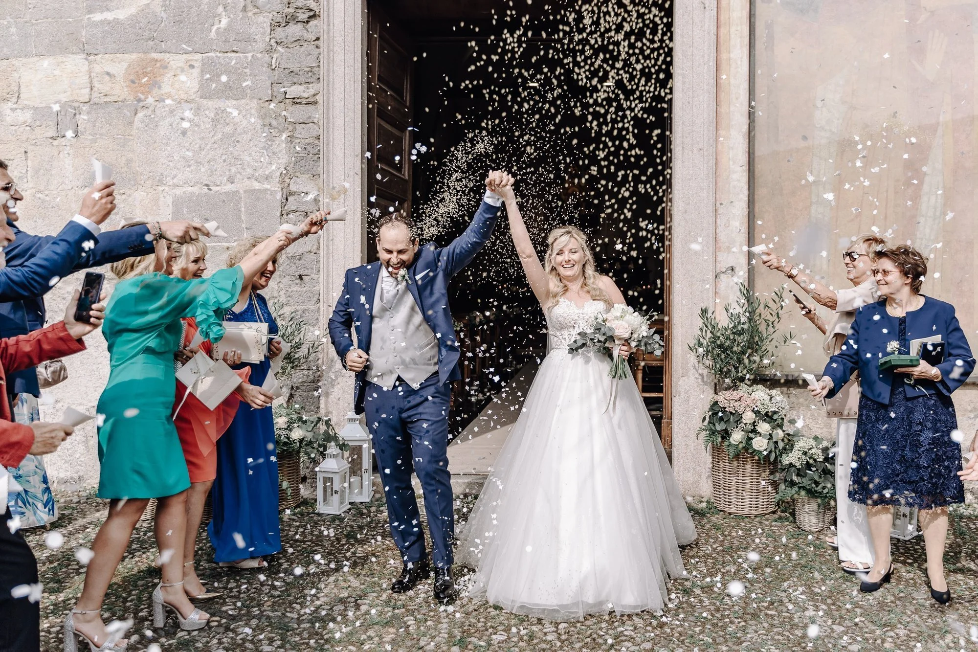 A newlywed couple celebrating outside a church with friends and family throwing confetti, the bride holding a bouquet and smiling, and the groom raising their joined hands.