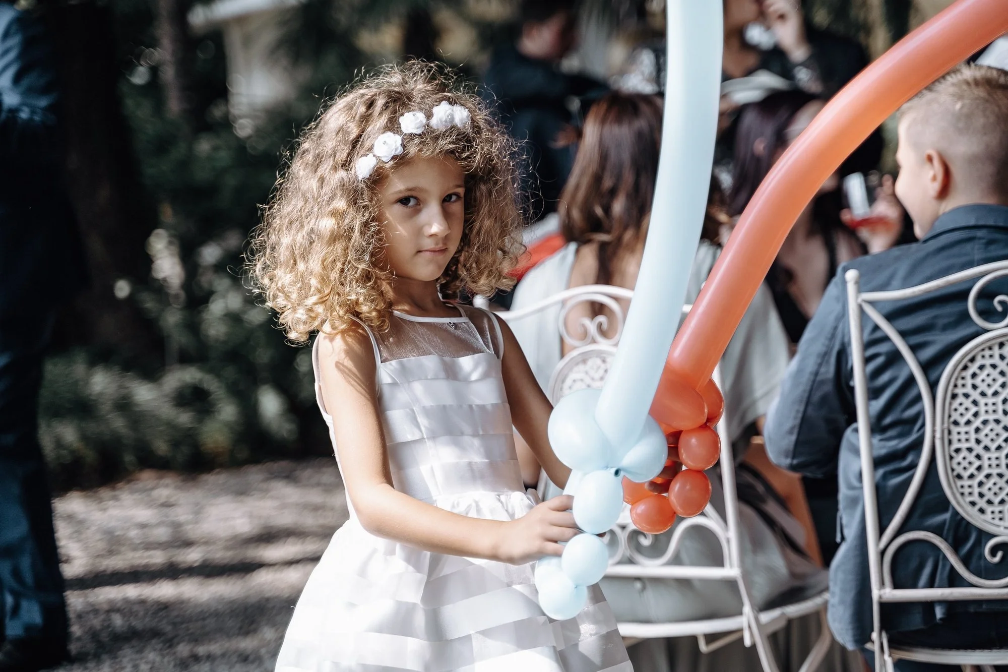 Young girl with curly hair wearing a white dress and floral headband, holding balloon animals, sitting on a white chair at an outdoor event with people in the background.