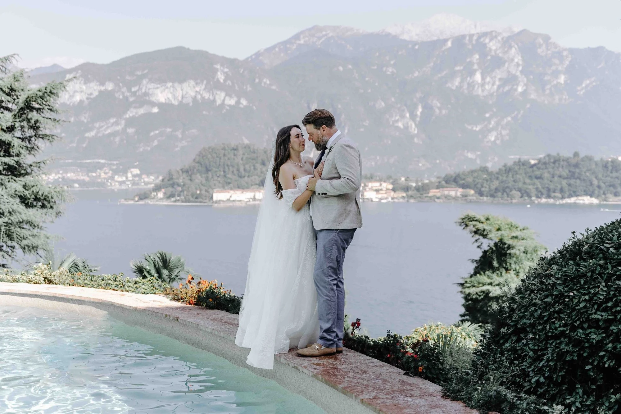 A bride and groom standing by a pool with mountains and a lake in the background during their wedding photoshoot.