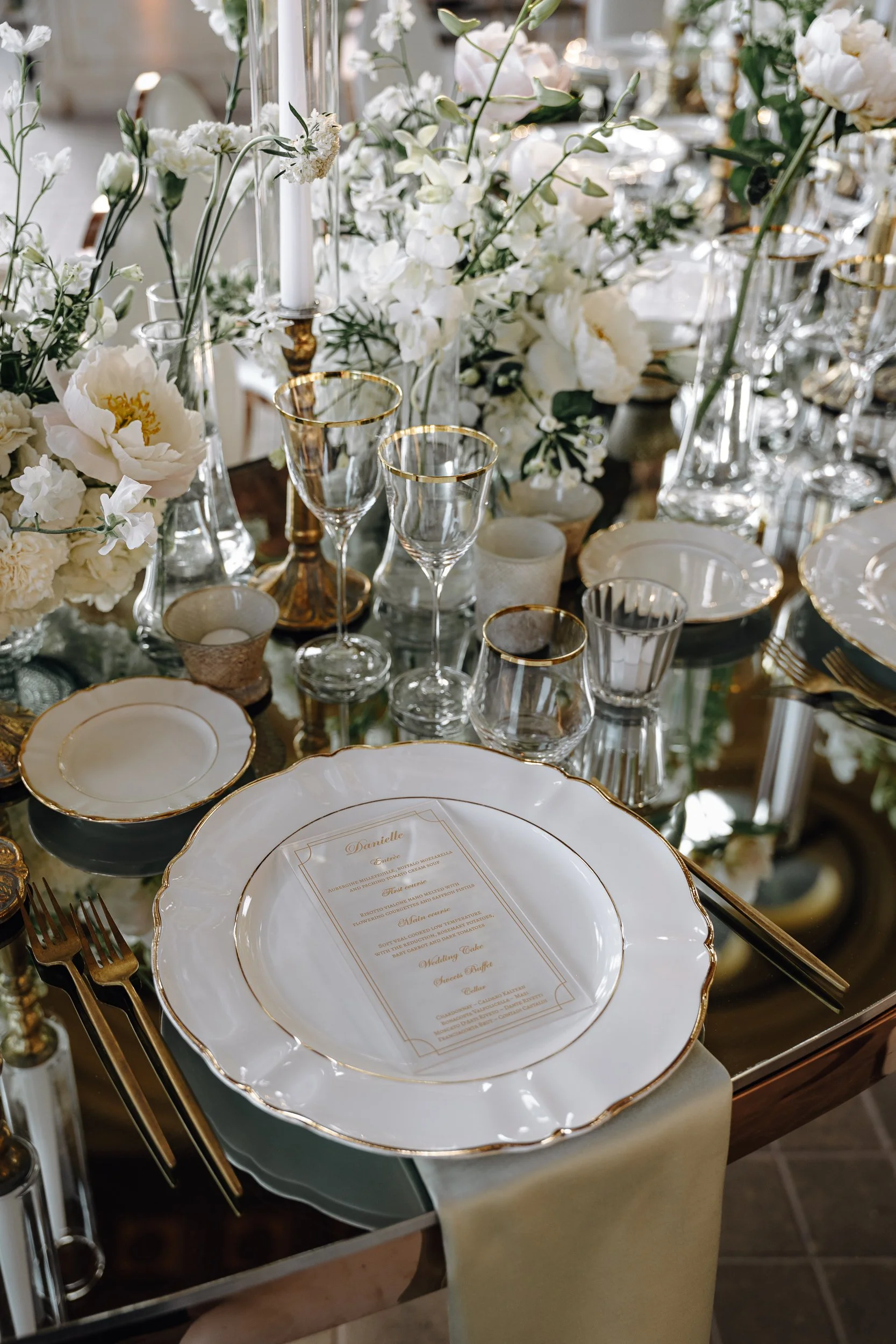 Elegant table setting with white and gold-rimmed china plates, gold cutlery, and various glassware including wine glasses and candle holders. The table is decorated with lush white flowers and tall candles, creating a sophisticated atmosphere.