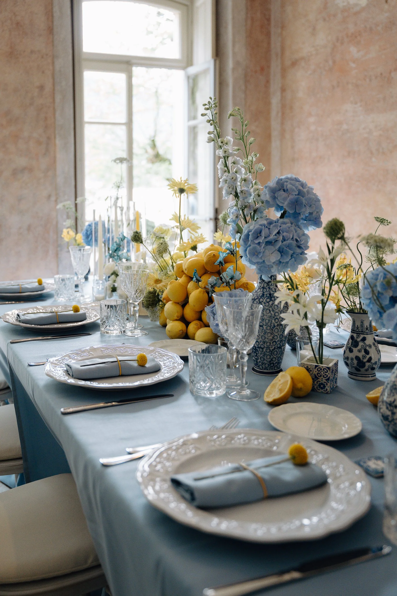 A dining table decorated with blue and white ceramics, fresh flowers, lemons, and clear glassware, set near a window with natural light.